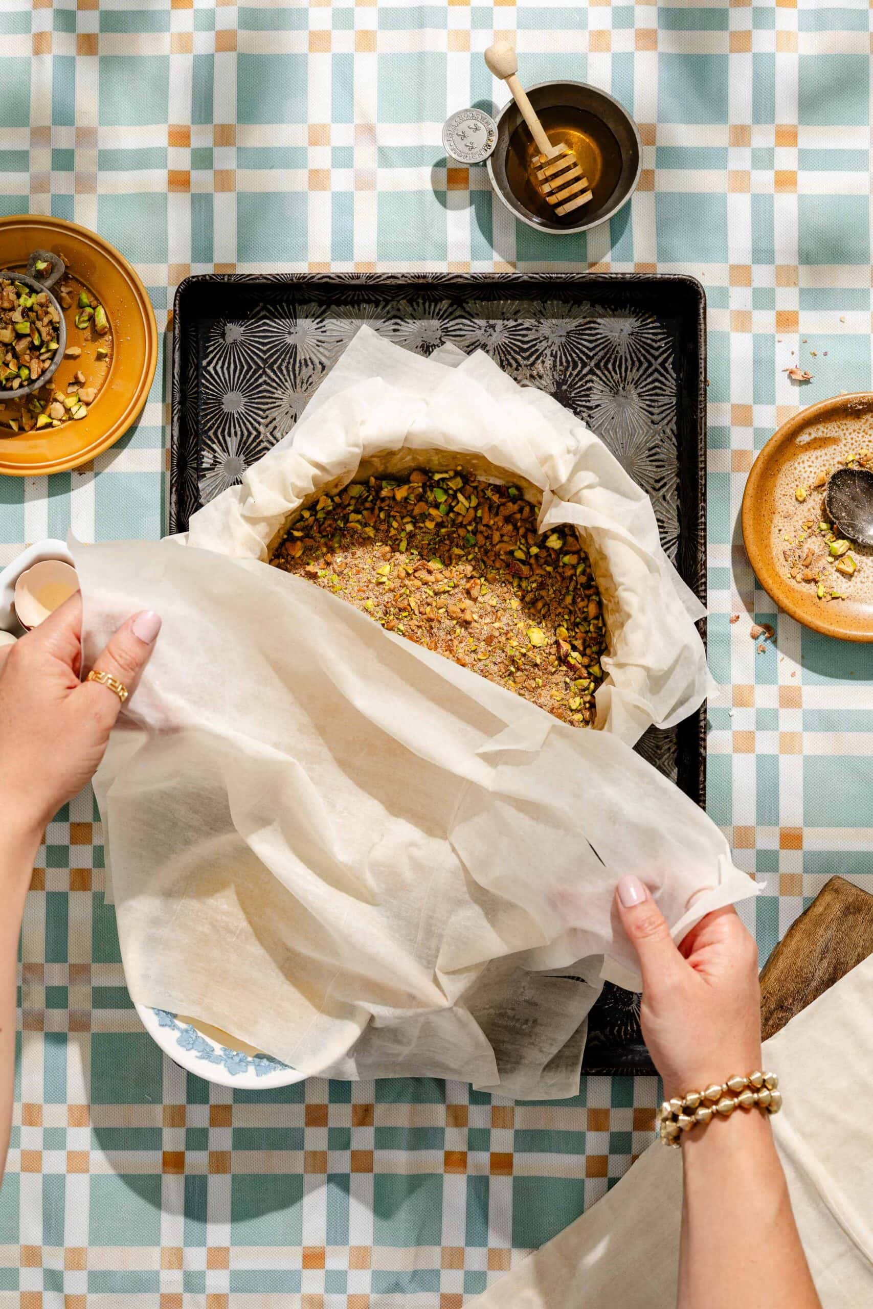 Hands wrapping a round pastry with parchment paper on a baking tray, topped with chopped nuts. Surrounding dishes hold honey, nuts, and seeds on a patterned tablecloth.