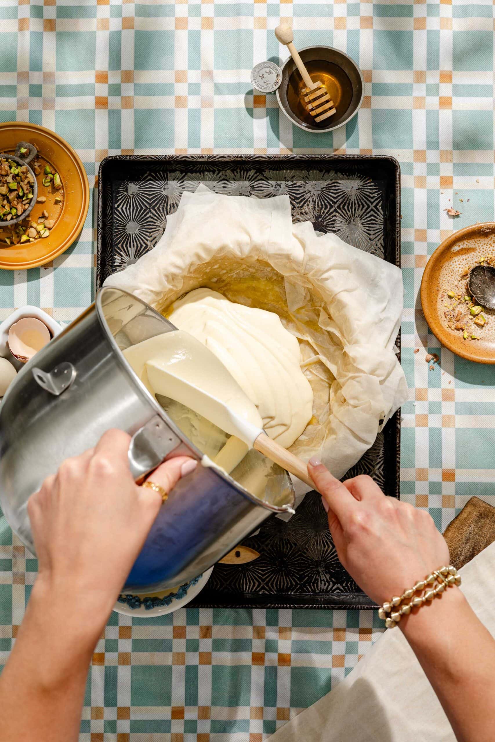 A person pours cake batter from a mixing bowl into a parchment-lined pan on a baking tray, surrounded by bowls of ingredients on a checkered tablecloth.