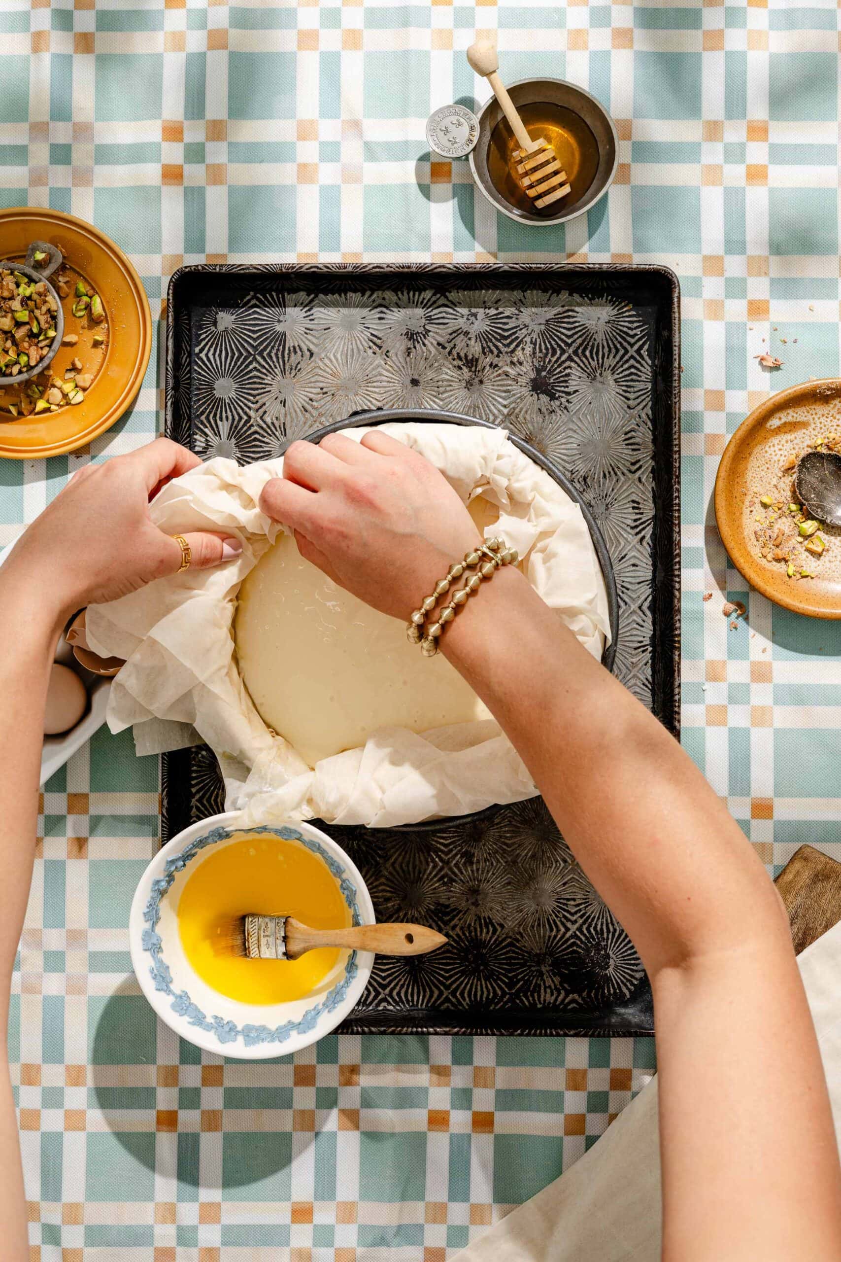 A person folds pastry dough into a round pan on a baking tray, with bowls of honey, nuts, and melted butter nearby on a checkered tablecloth.