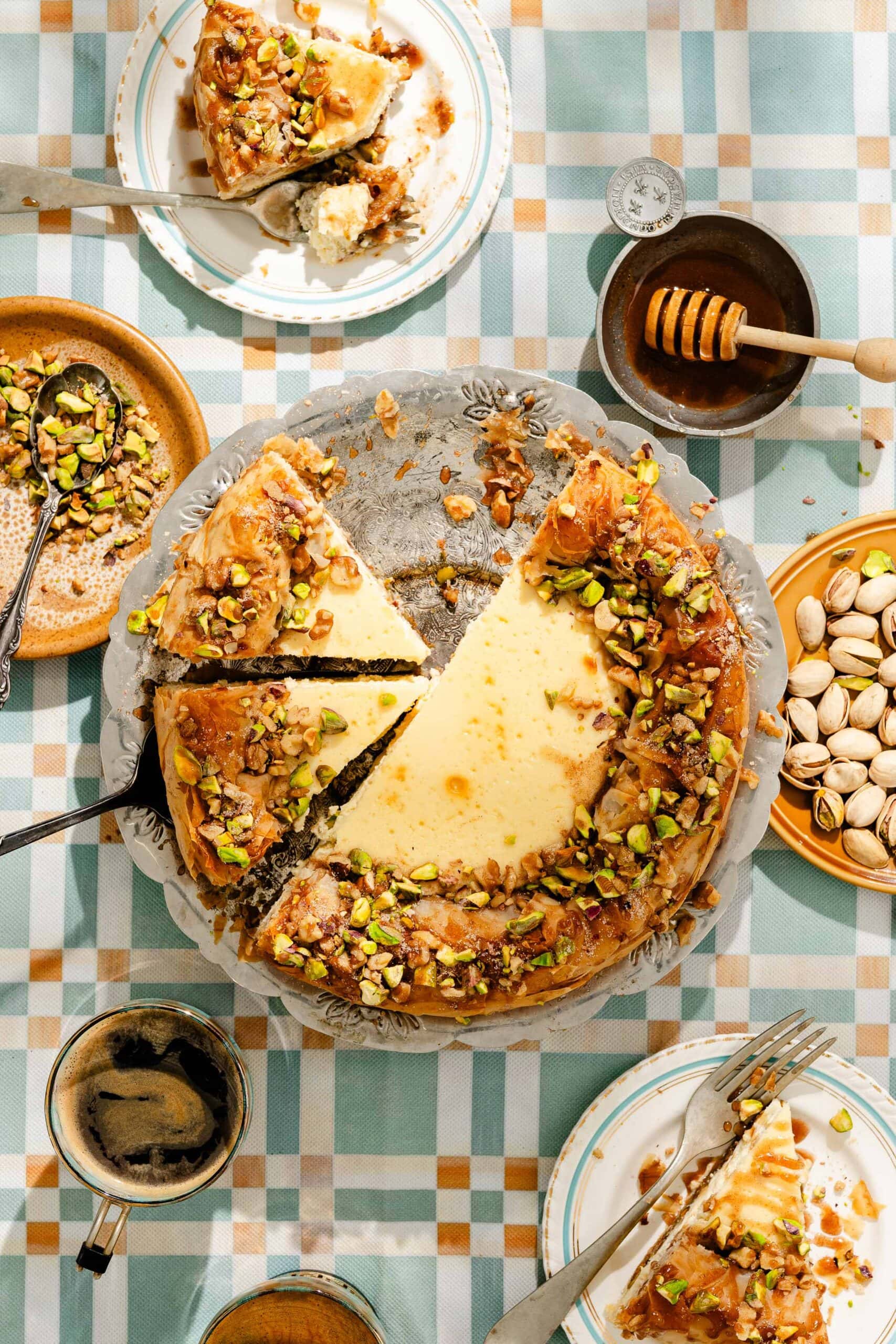 A cheesecake topped with chopped pistachios sits on a table with slices served on plates, cups of coffee, a bowl of pistachios, honey dipper, and utensils, all on a checkered tablecloth.