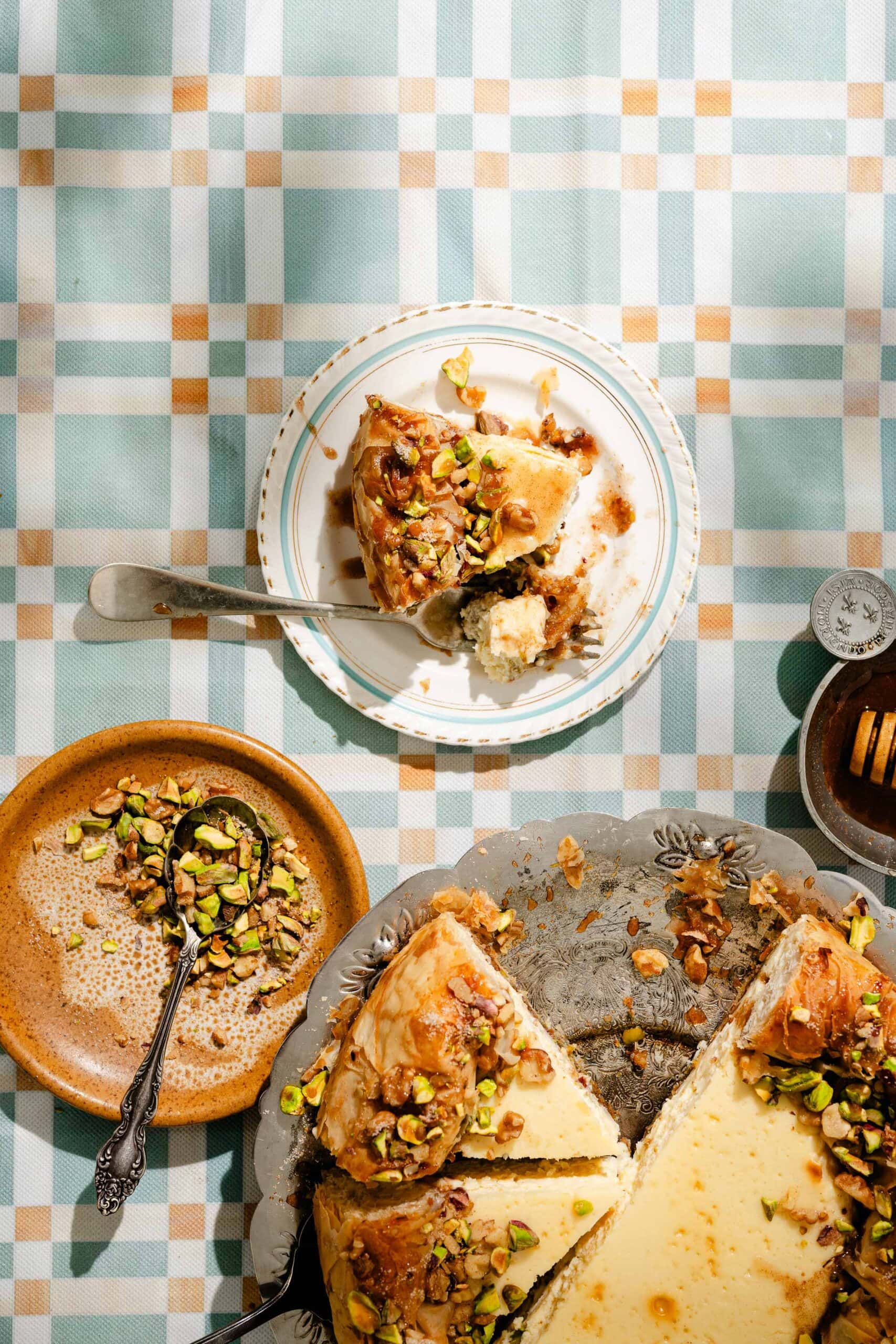 A slice of cheesecake topped with chopped pistachios sits on a plate with a fork. Nearby are a partial cheesecake, a bowl of pistachios, and a jar of honey on a blue and beige plaid tablecloth.