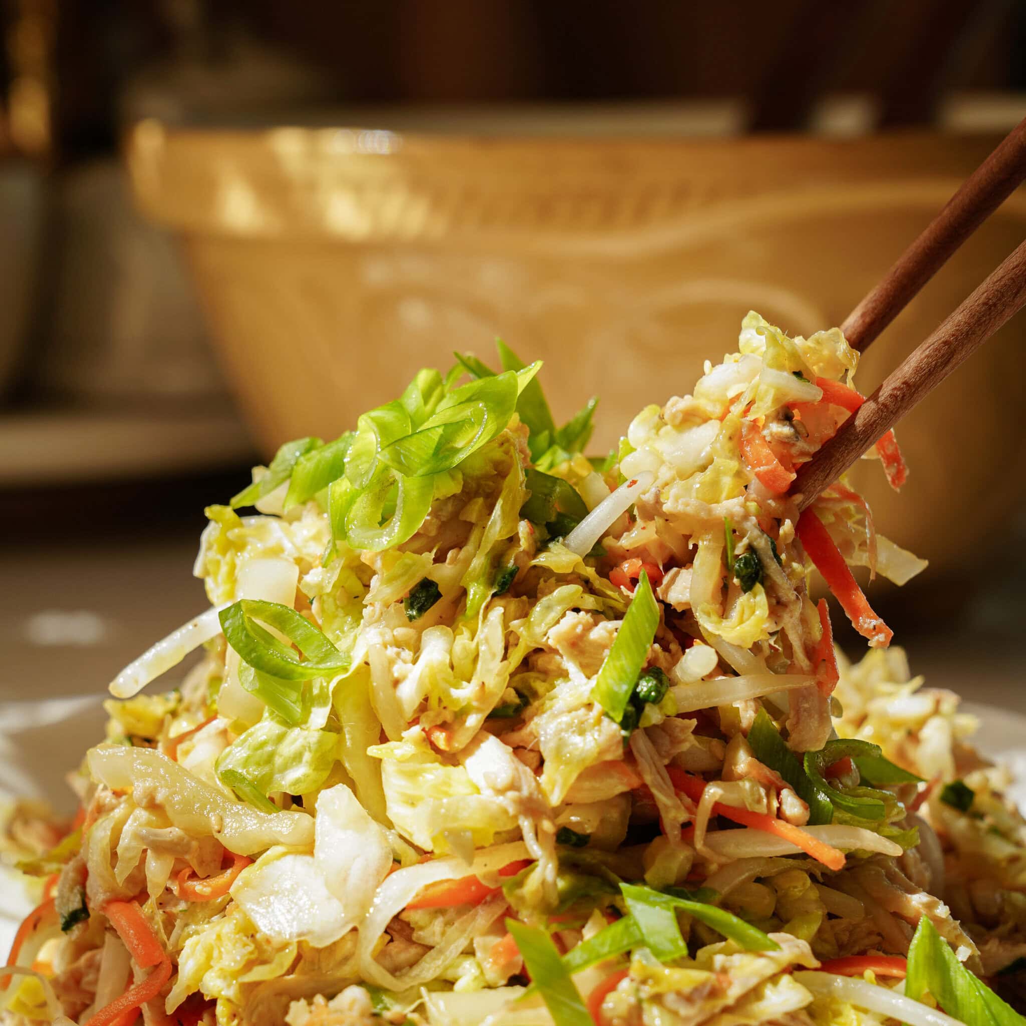 Chopsticks holding up a colorful Asian salad made of shredded cabbage, carrots, green onions, and fresh herbs, with a bowl blurred in the background.