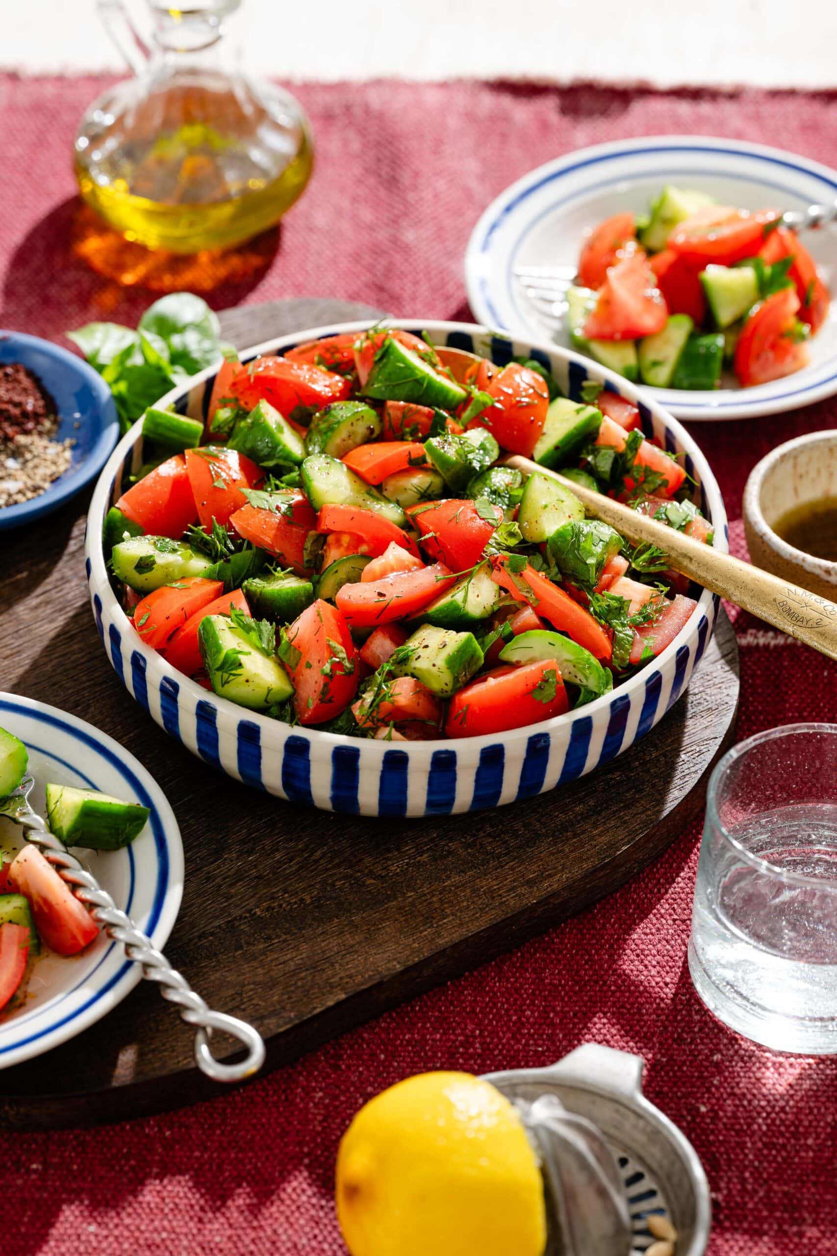 A bowl of chopped tomato and cucumber salad with herbs, surrounded by small plates of salad, a glass of water, olive oil, a bowl of spices, and a lemon on a red tablecloth.