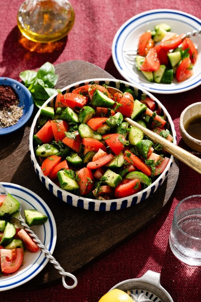 A bowl of fresh salad with chopped tomatoes, cucumbers, and herbs on a wooden board, surrounded by small plates of salad, seasonings, a glass of water, and a bottle of olive oil on a red tablecloth.
