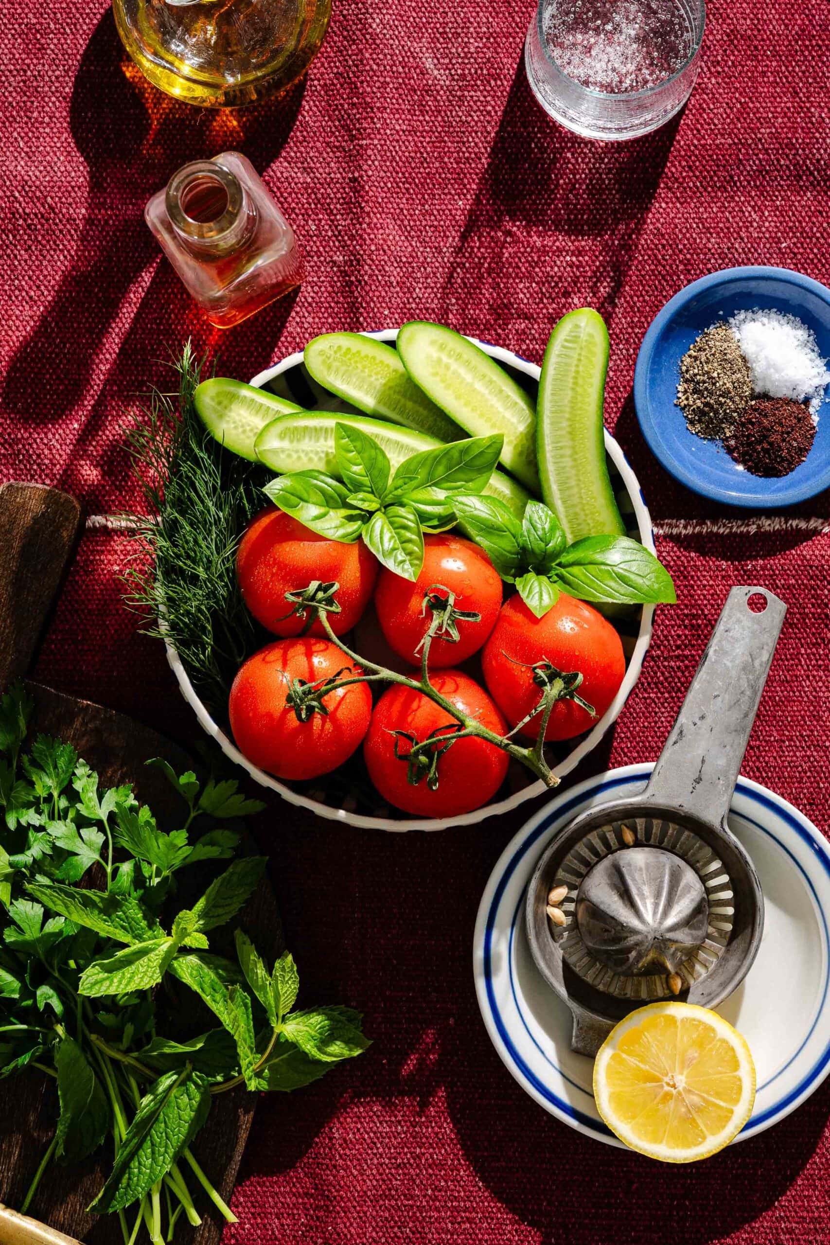 A bowl of tomatoes, cucumbers, basil, and dill on a red cloth, surrounded by fresh herbs, a lemon half, a citrus juicer, oil, and bowls of salt and pepper.