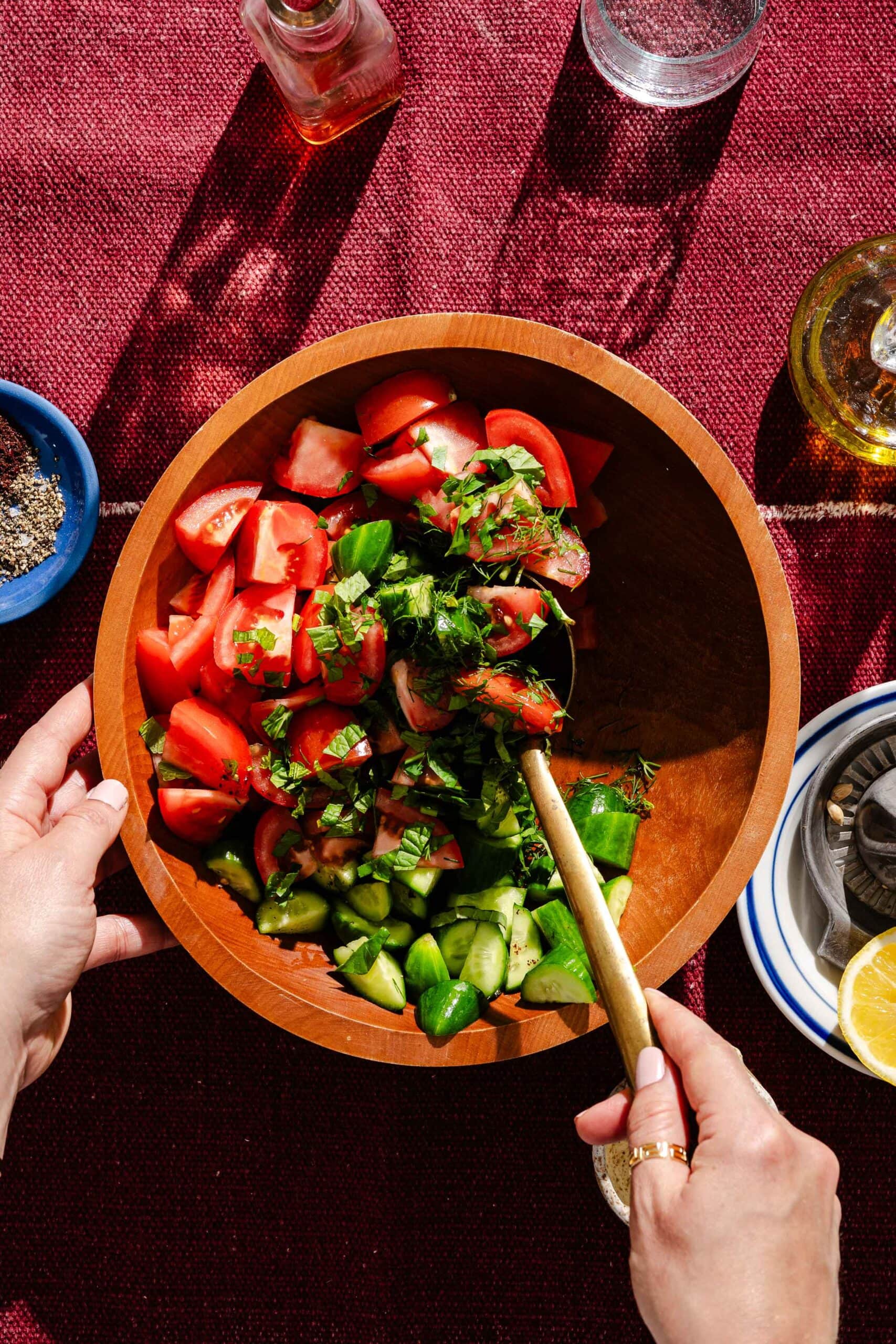 A person mixes a fresh vegetable salad with tomatoes, cucumbers, and herbs in a wooden bowl on a red tablecloth, surrounded by seasonings, oil, and lemon.
