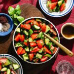 A bowl of fresh salad with chopped tomatoes, cucumbers, herbs, and a wooden spoon. Small plates of salad, seasonings, and a glass of water are nearby on a red tablecloth.