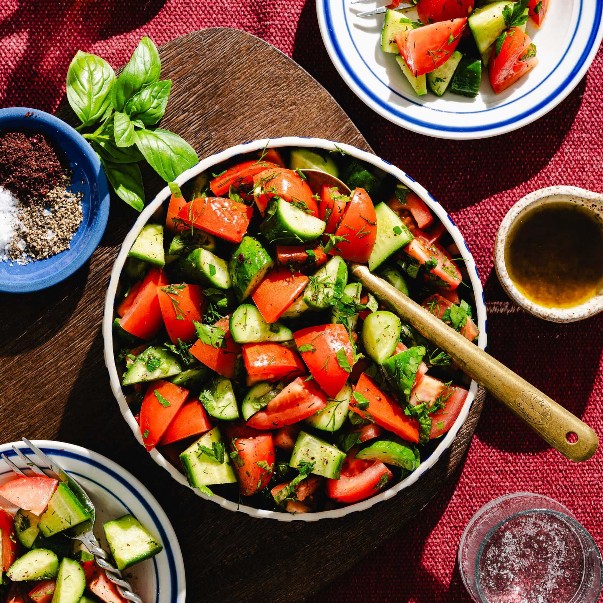 A bowl of fresh salad with chopped tomatoes, cucumbers, herbs, and a wooden spoon. Small plates of salad, seasonings, and a glass of water are nearby on a red tablecloth.