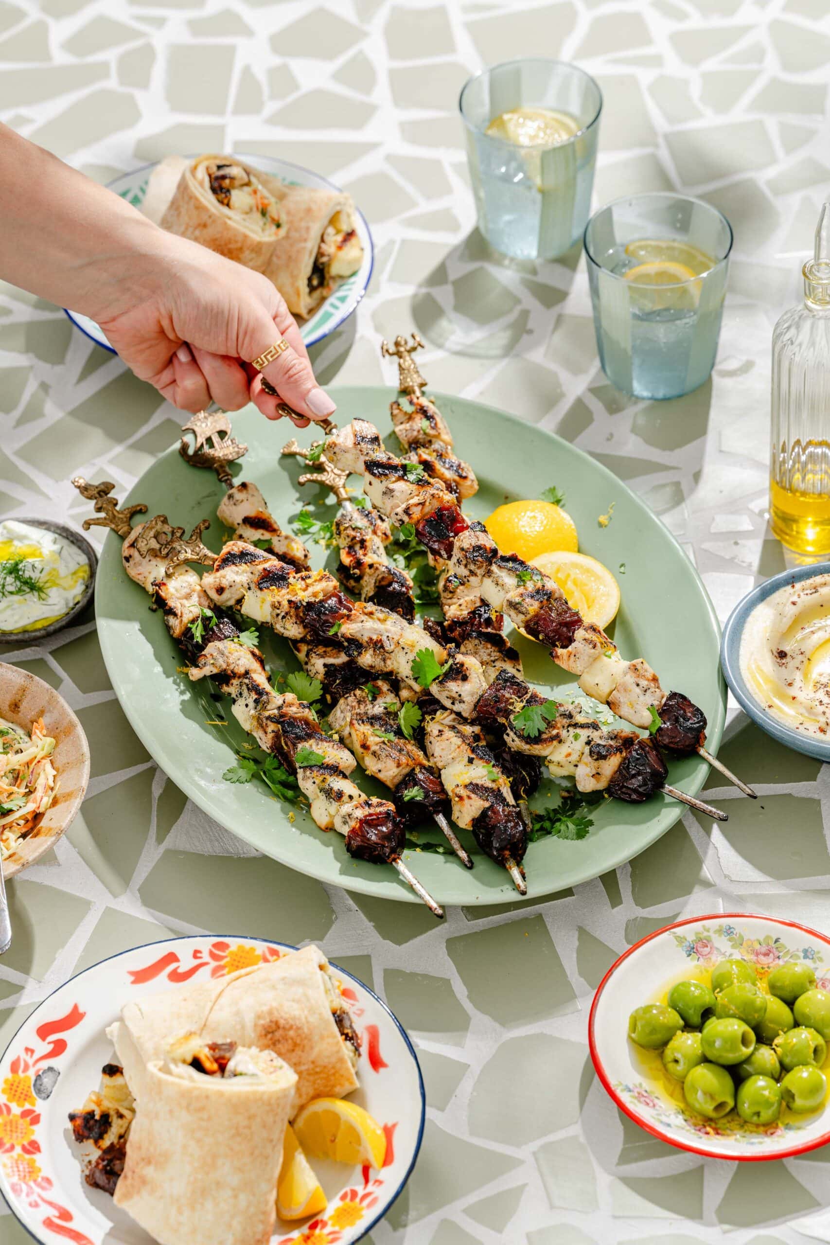 A hand picks up a grilled chicken and vegetable skewer from a platter, surrounded by dishes of olives, pita wraps, dips, and drinks on a patterned table.