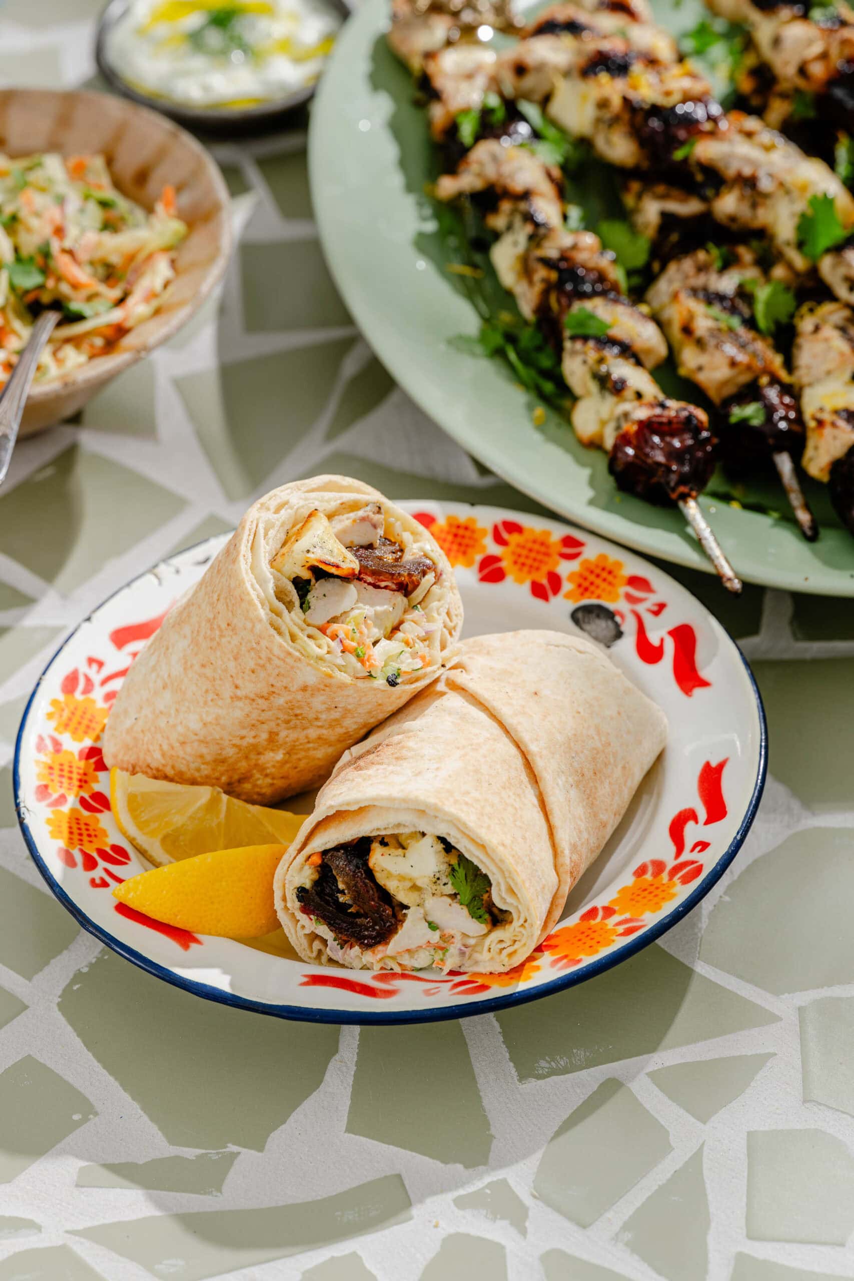 A plate with two halves of a wrap filled with grilled meat and vegetables, garnished with lemon wedges. In the background, skewers of grilled meat are on a green platter.