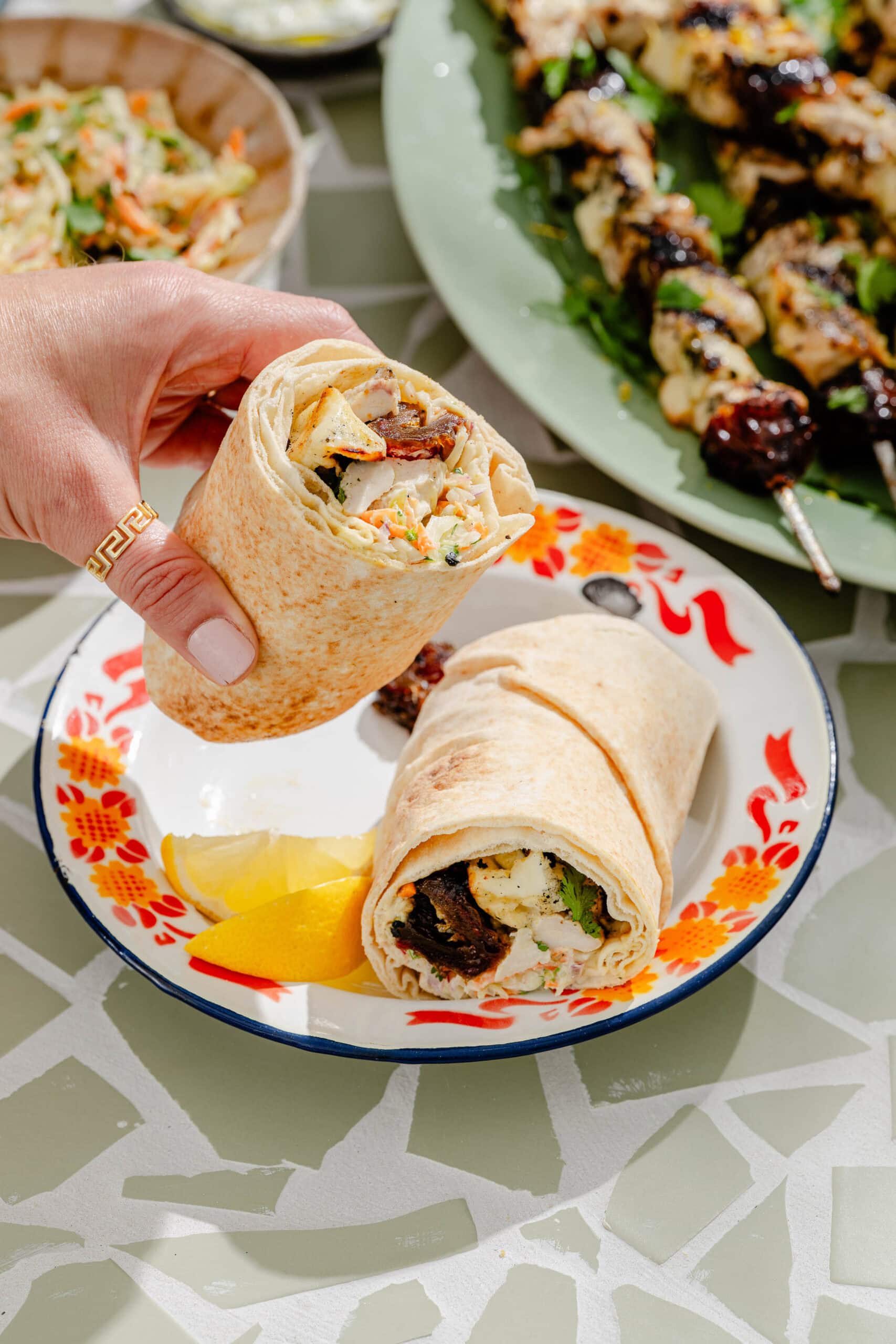 A hand holds half of a wrap filled with grilled meat and vegetables over a plate with another wrap and lemon wedges. Skewers of grilled meat and vegetables are on a green platter nearby.