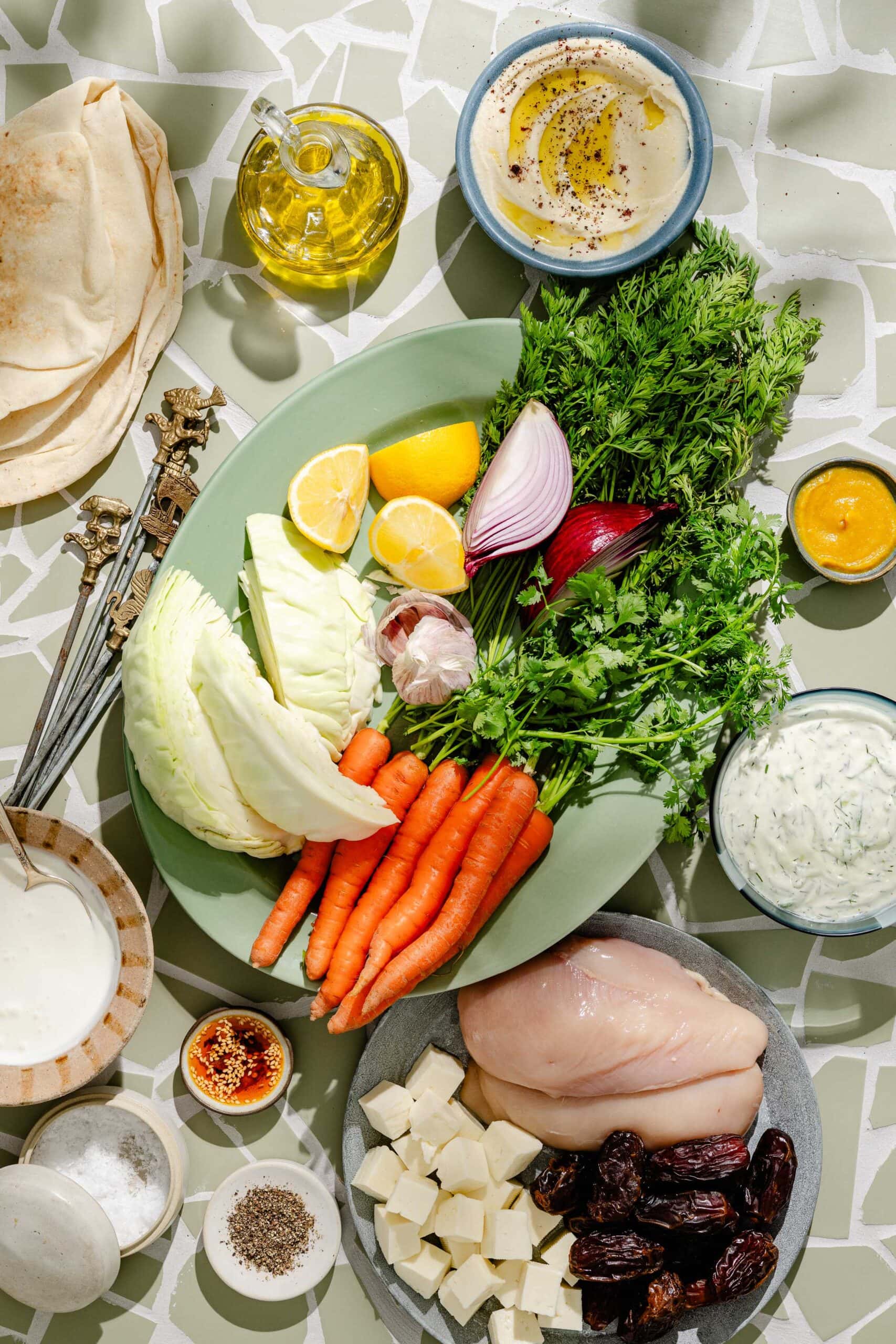 A table with raw chicken, feta, dates, carrots, cabbage, herbs, onion, lemon, flatbread, olive oil, hummus, tzatziki, yogurt, spices, and metal skewers arranged on a mosaic-patterned surface.