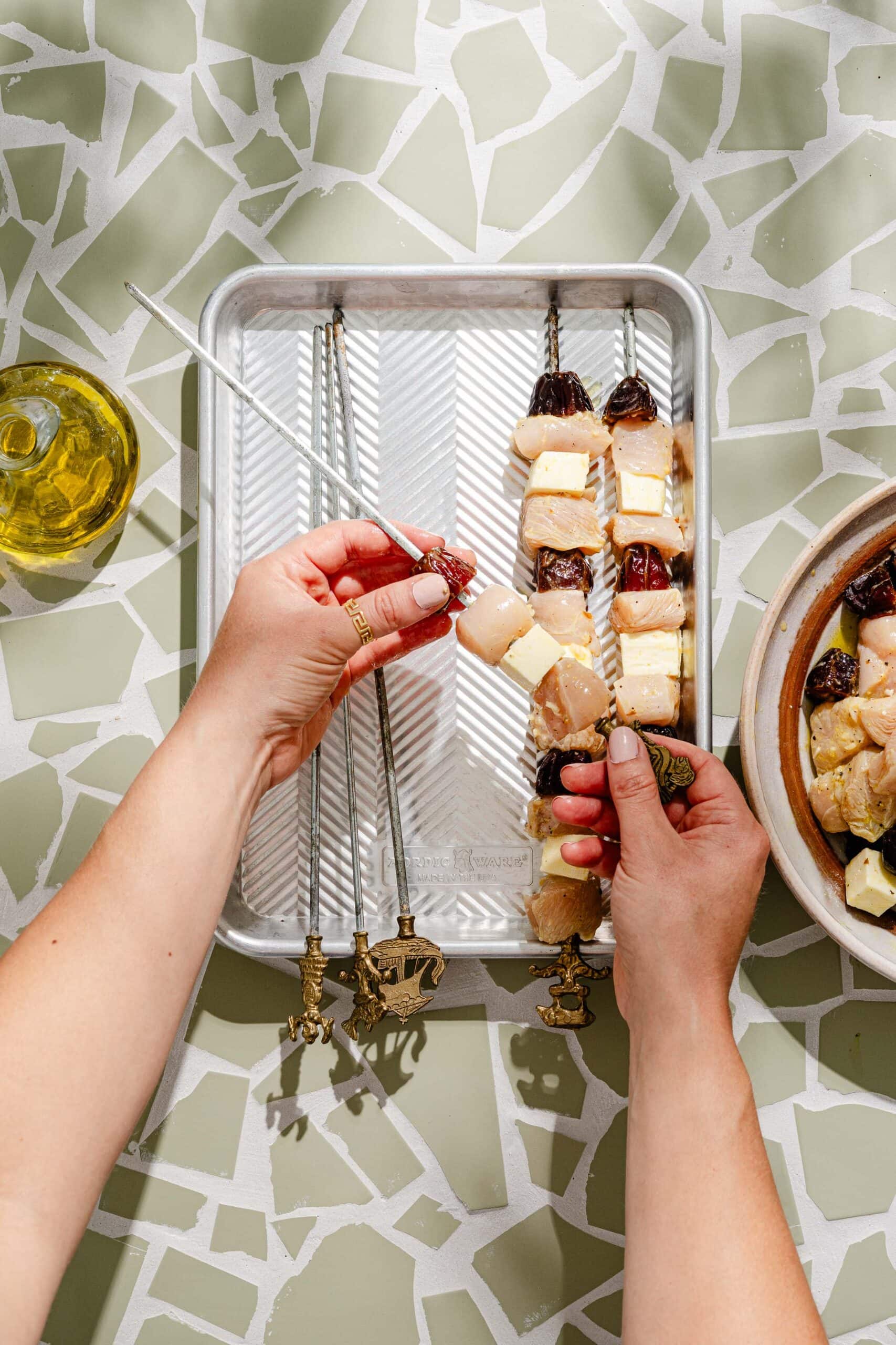 A person threads skewers with cheese, dates, and what appears to be scallops on a baking tray. There is a bowl of ingredients and a bottle of oil nearby on a green and white patterned surface.