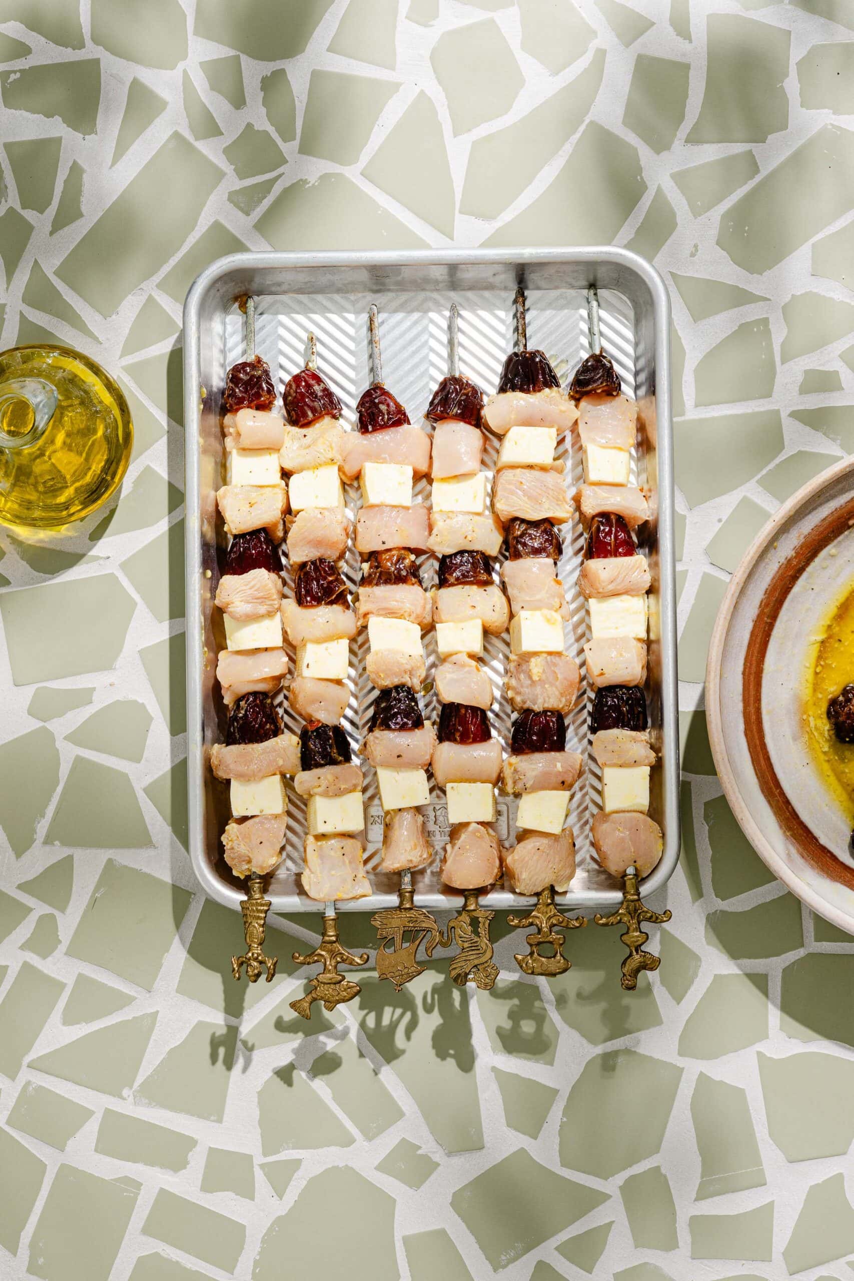 Metal skewers in a pan, each with cubes of chicken, cheese, and dark dried fruit, arranged in rows. The pan sits on a patterned white and light green surface. A bottle and bowl are nearby.
