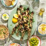 Grilled chicken skewers on a green platter with lemon wedges, surrounded by olives, hummus, salad, yogurt dip, and flatbread on a patterned table.