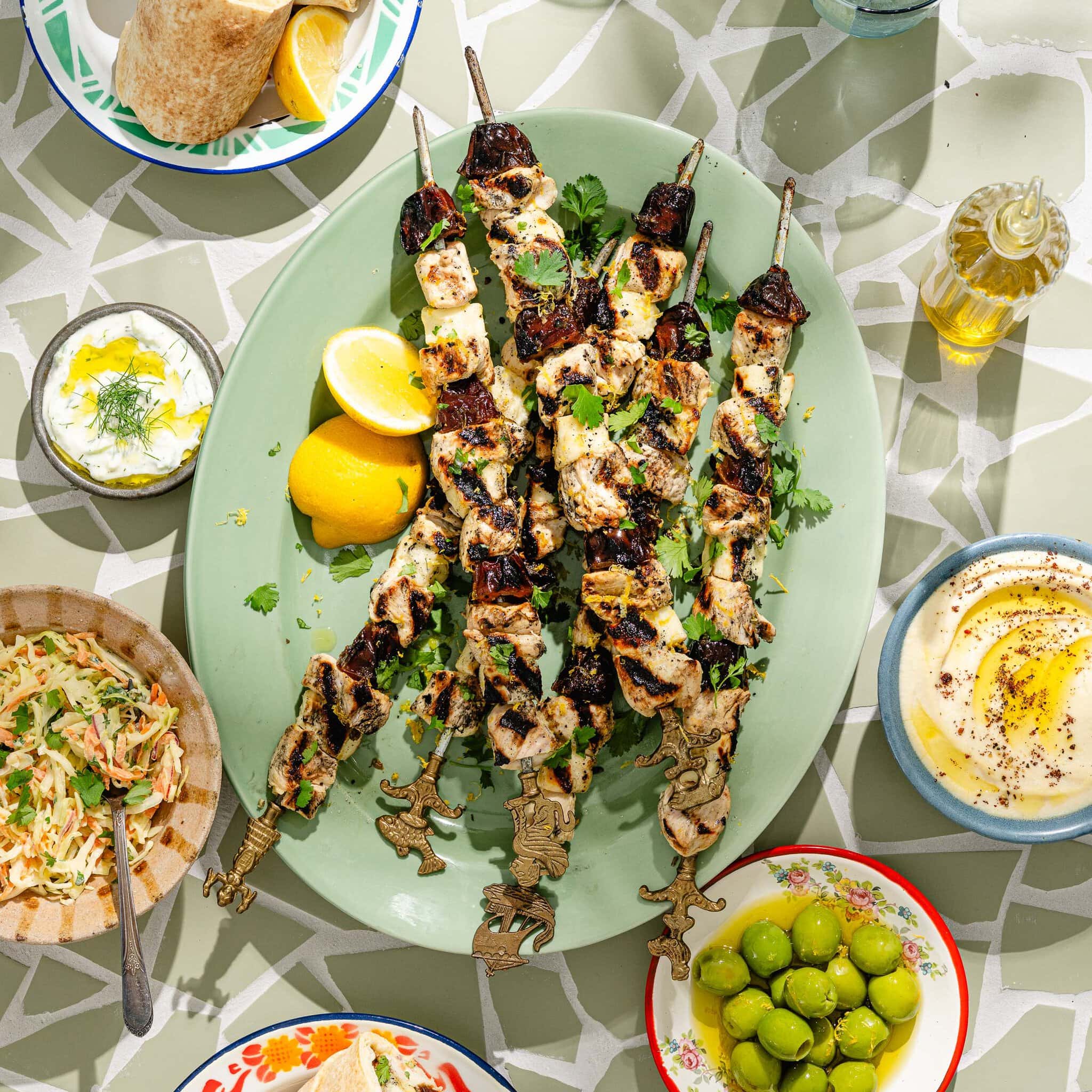 Grilled chicken skewers on a green platter with lemon wedges, surrounded by olives, hummus, salad, yogurt dip, and flatbread on a patterned table.