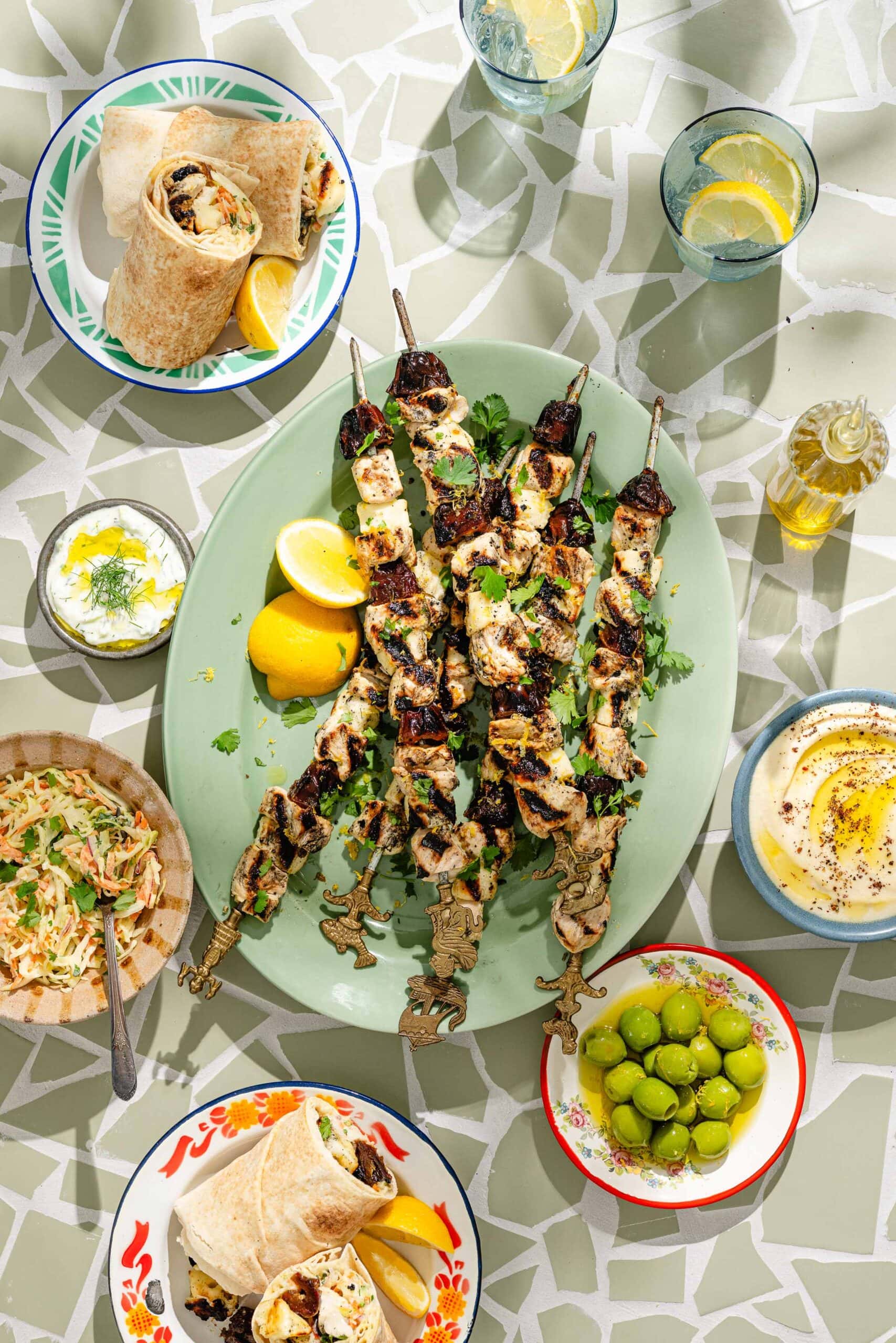 A table set with chicken skewers, lemon wedges, hummus, green olives, tzatziki, salad, and wraps, with glasses of water and lemon on a tiled surface.
