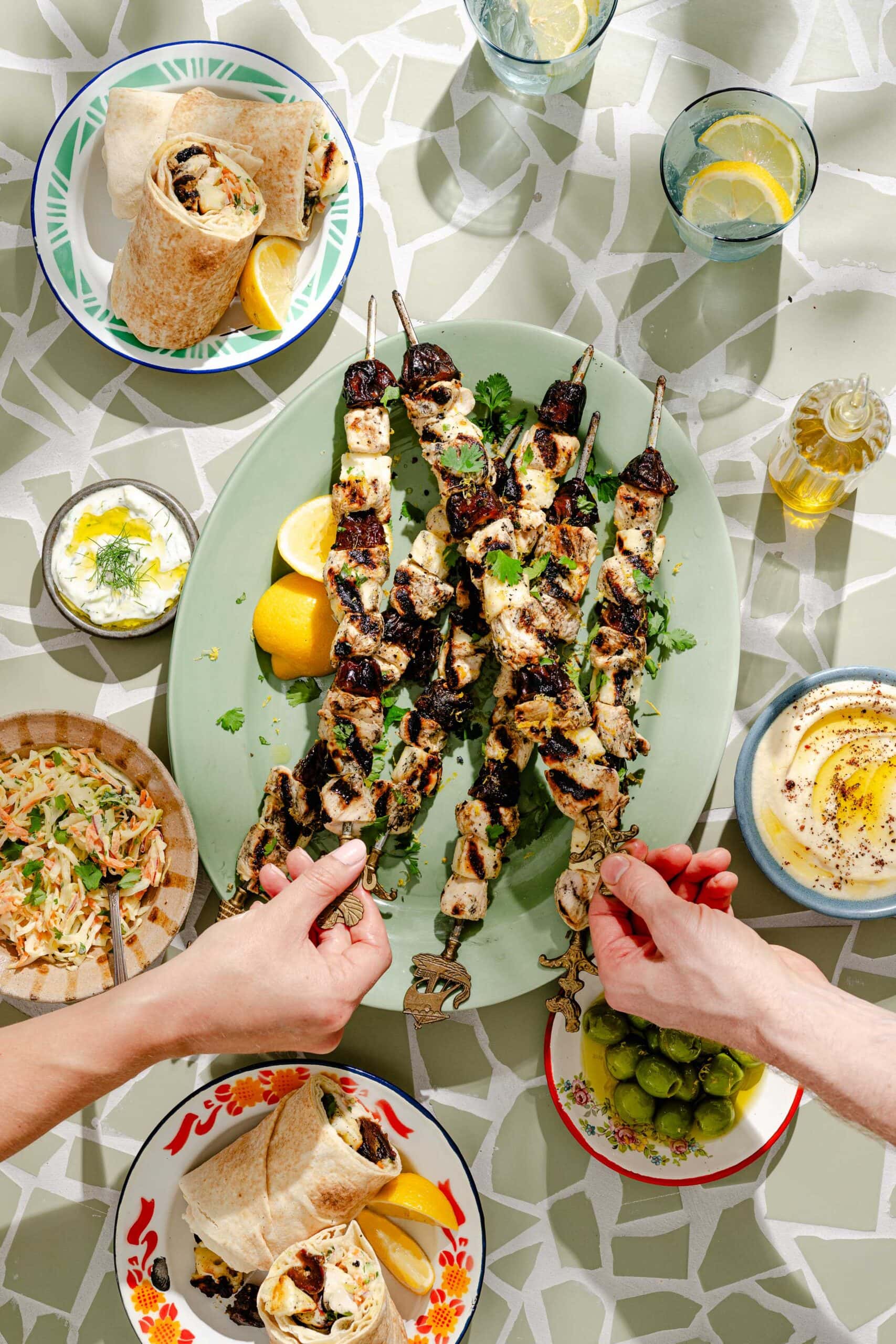 Hands reaching for grilled chicken kebabs on a platter with lemon, surrounded by pita wraps, olives, salad, dip, and drinks on a patterned table. Sunlight casts bright shadows on the scene.