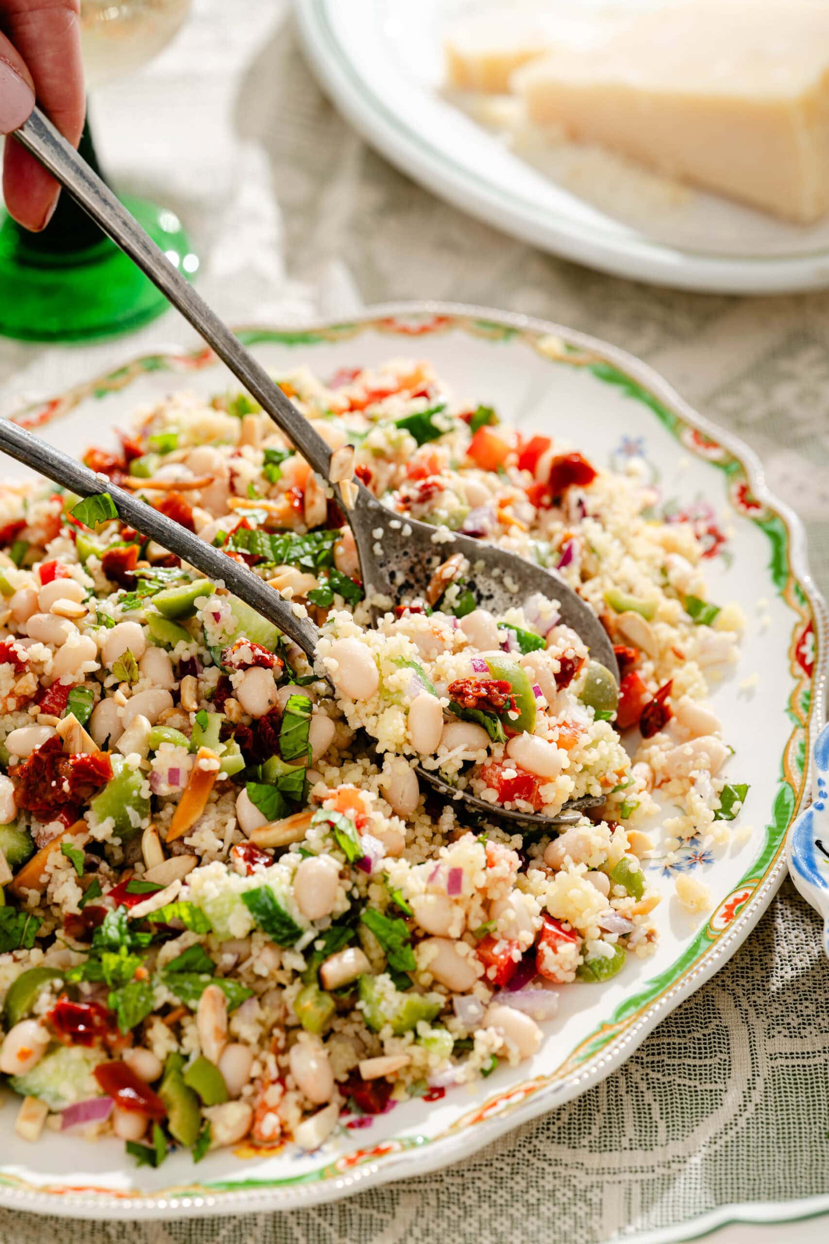 A close-up of a colorful bean and couscous salad being served with utensils on a decorative plate, with visible chopped vegetables and herbs. A block of cheese sits on a plate in the background.