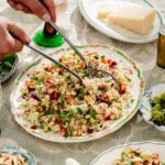 A person serves a colorful bean and vegetable couscous salad from a decorative plate on a lace tablecloth; cheese, green olives, and a wine glass are nearby.