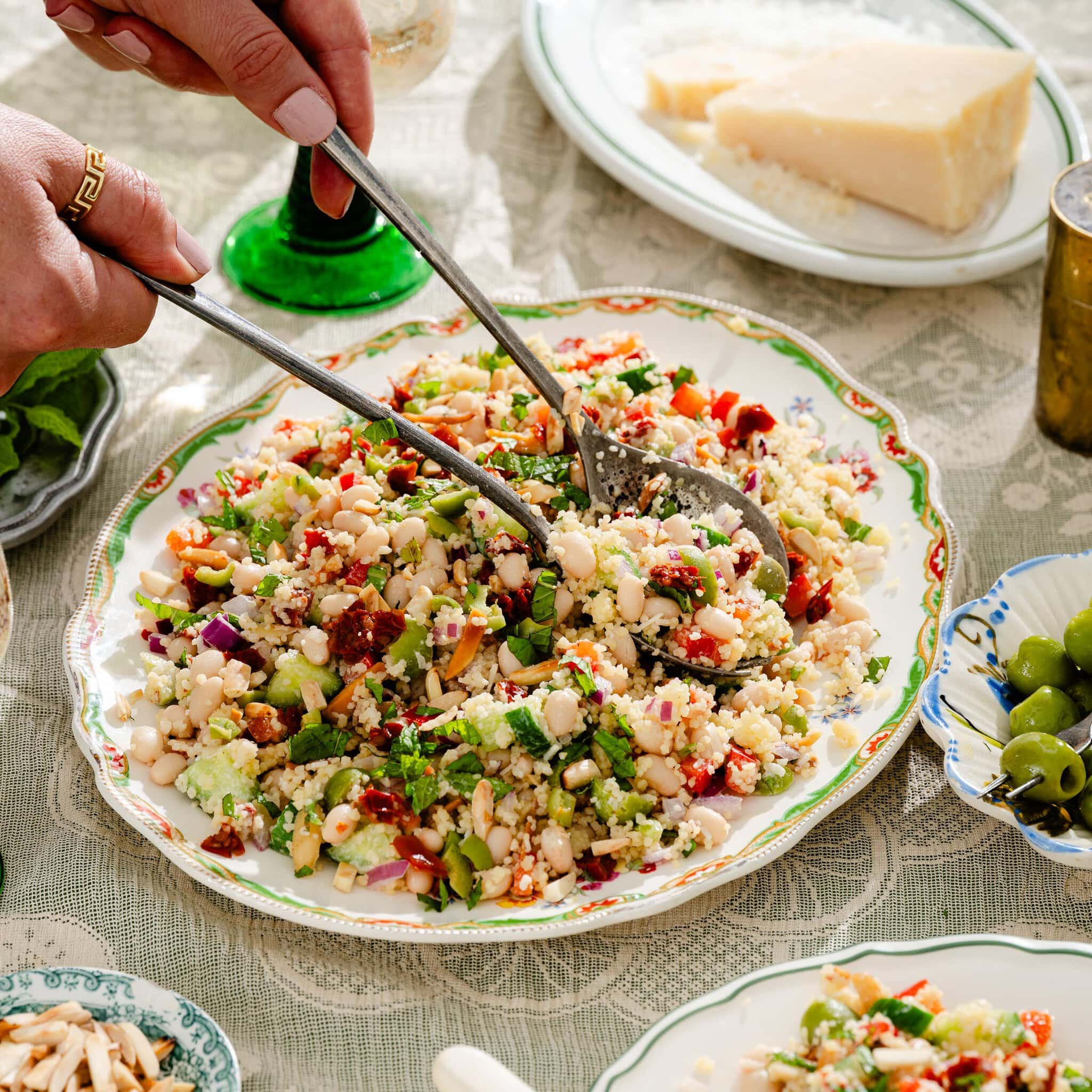 A person serves a colorful bean and vegetable couscous salad from a decorative plate on a lace tablecloth; cheese, green olives, and a wine glass are nearby.
