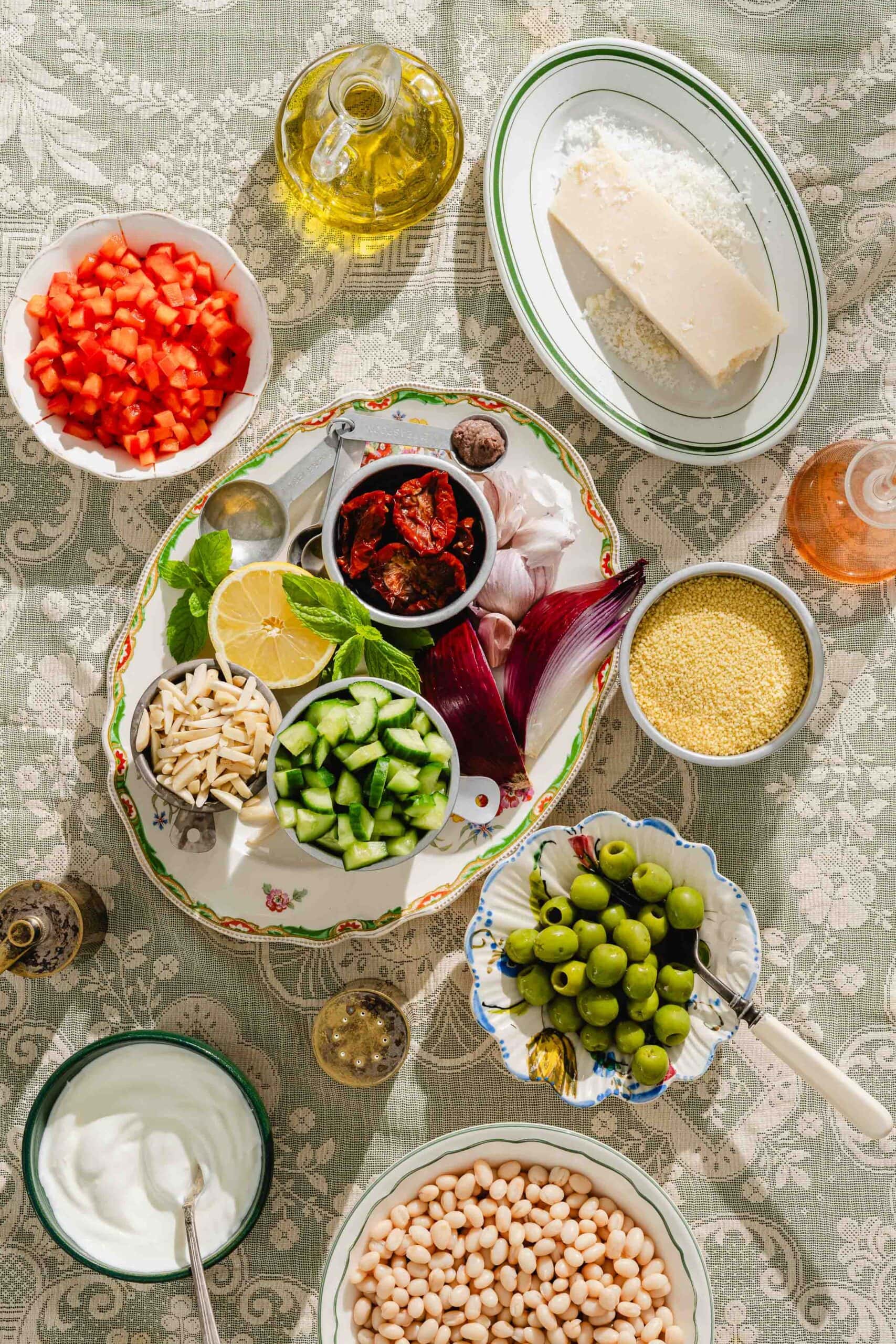 Assorted ingredients on a table, including diced red pepper, parmesan, couscous, cucumber, sun-dried tomatoes, red onion, lemon, fresh herbs, olives, white beans, yogurt, olive oil, and vinegar.