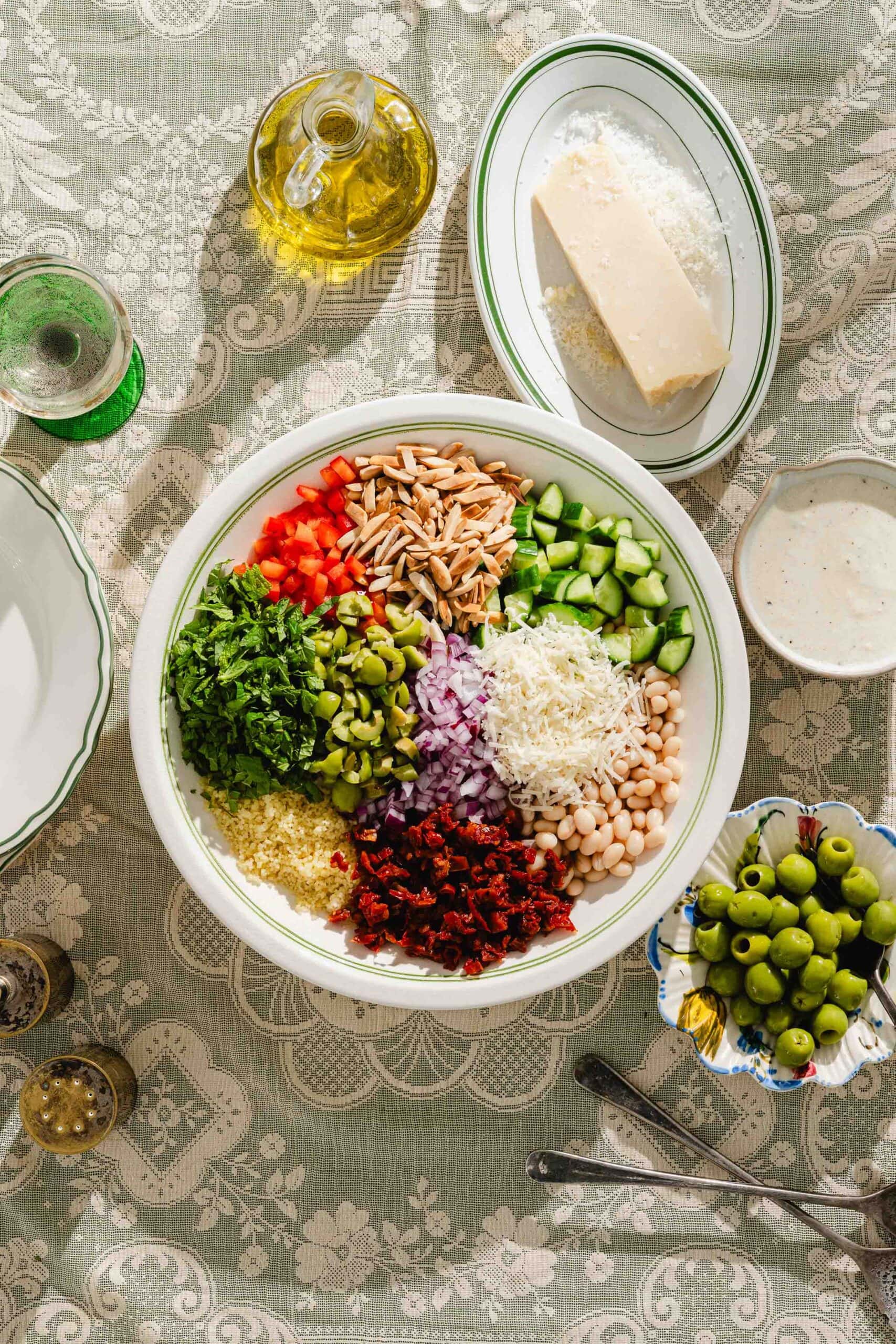 A bowl of colorful chopped vegetables, cheese, beans, and grains sits on a lace-covered table, surrounded by olive oil, parmesan cheese, a dish of green olives, and glassware.