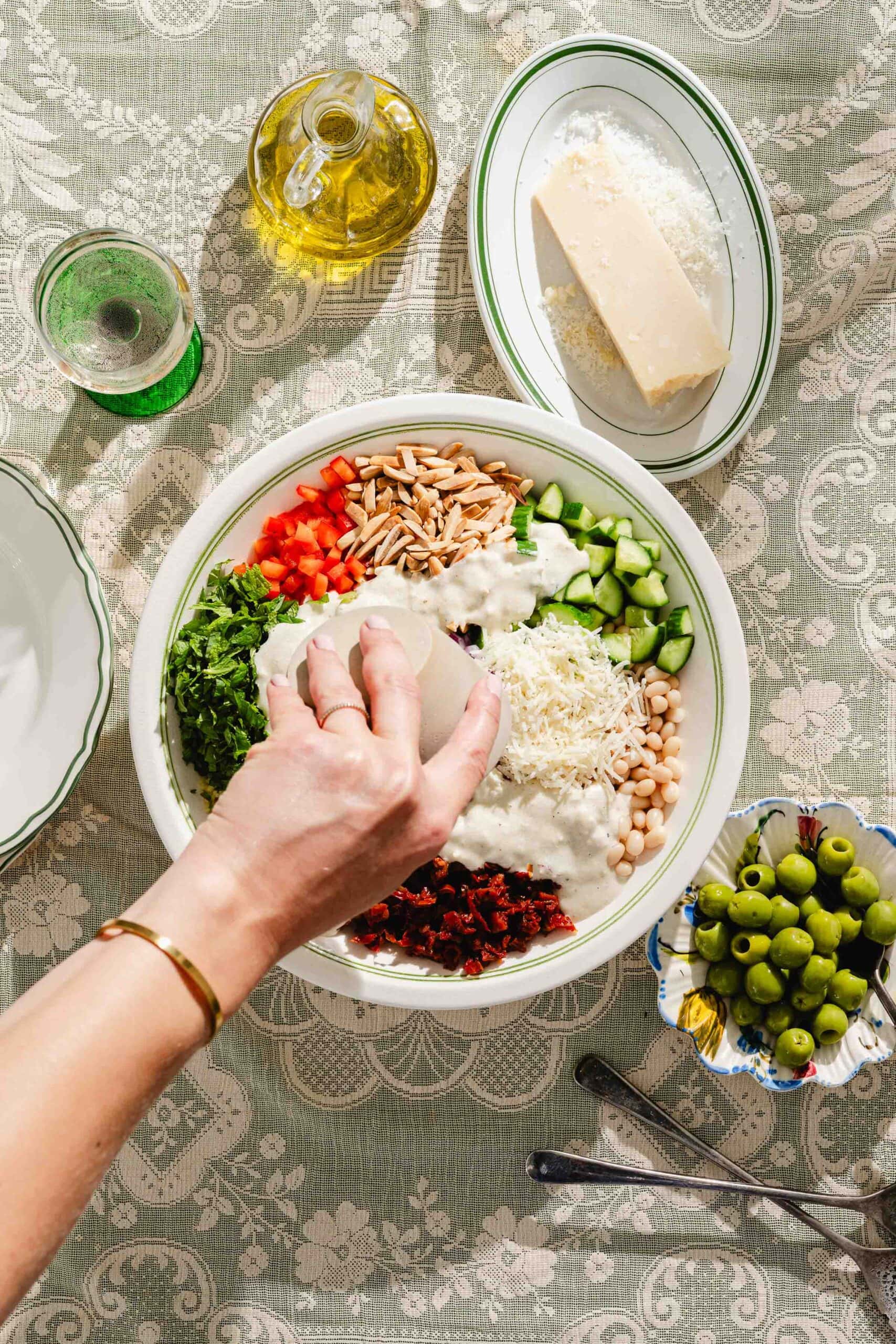 A hand adds dressing to a bowl of salad with vegetables, beans, grains, and cheese. Nearby are olives, grated cheese, olive oil, and a drink on a lace-patterned tablecloth.