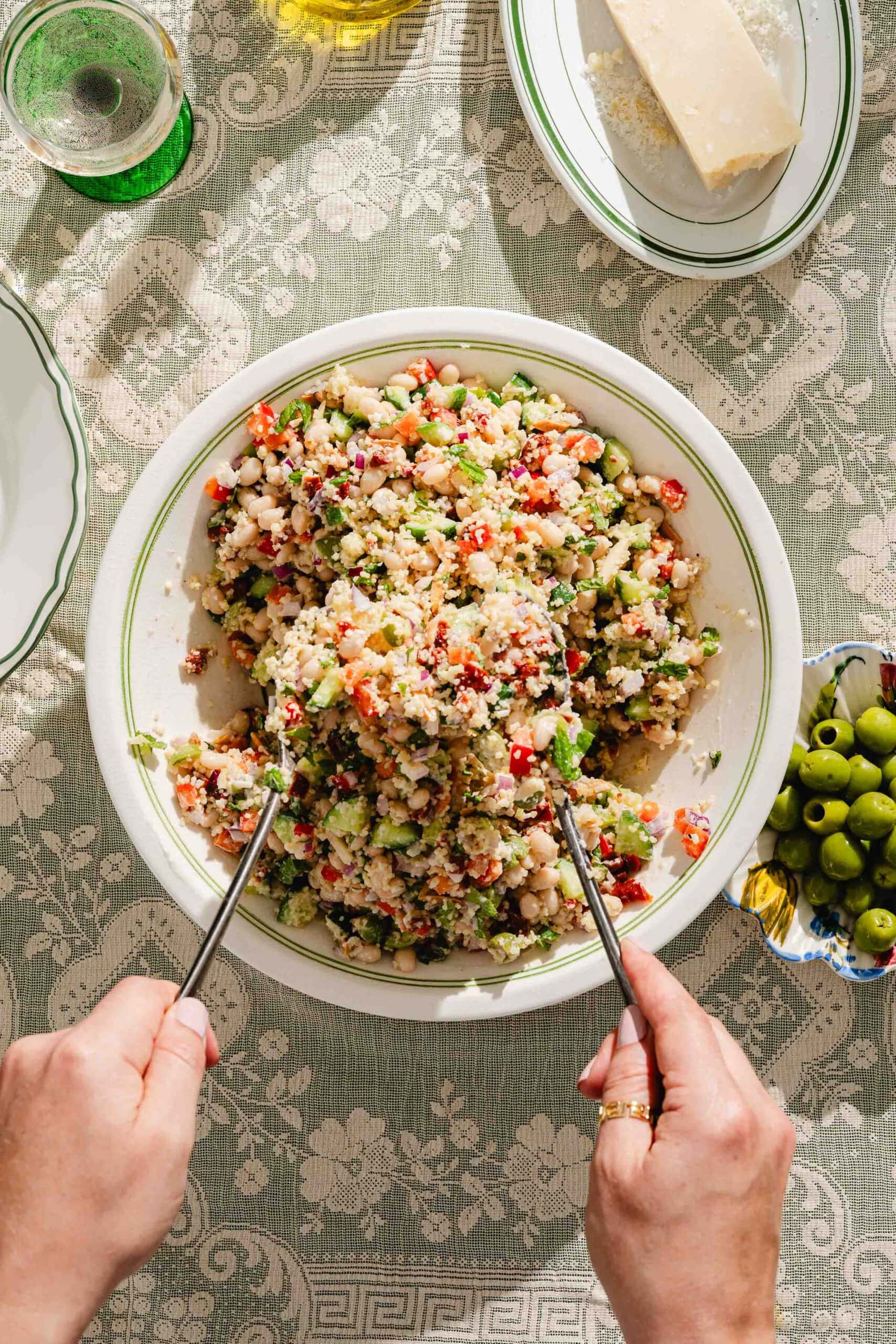 A person serves a colorful bean and vegetable salad from a large bowl on a patterned tablecloth, with green olives, a wedge of cheese, and a glass of water nearby.