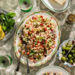A platter of colorful couscous salad with vegetables and herbs sits on a lace-covered table, surrounded by green olives, mint, lemon slices, a wedge of cheese, and two glasses of water.