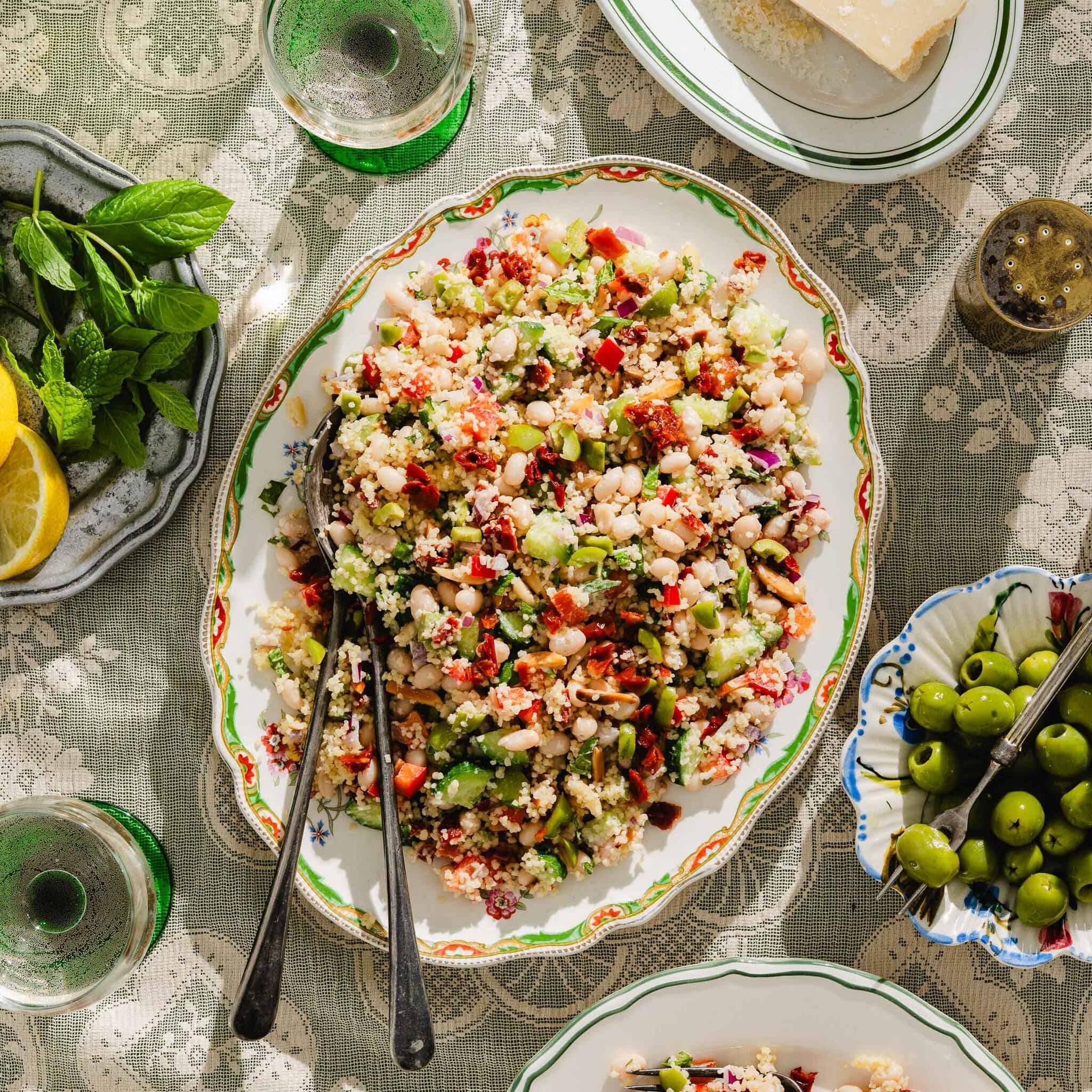 A platter of colorful couscous salad with vegetables and herbs sits on a lace-covered table, surrounded by green olives, mint, lemon slices, a wedge of cheese, and two glasses of water.
