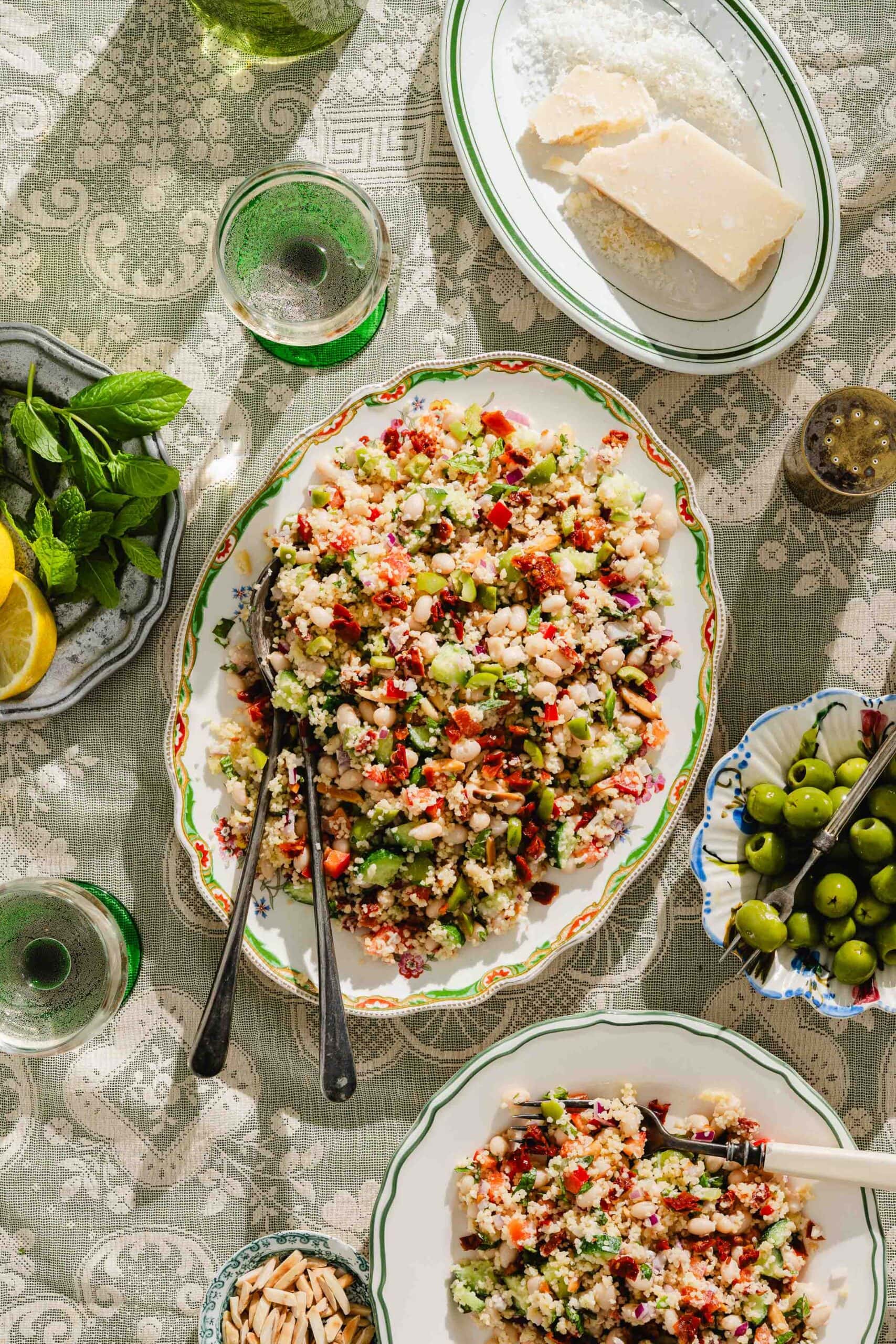A table set with a platter of couscous salad with vegetables, surrounded by plates of almonds, olives, lemon, mint, Parmesan cheese, and glasses of water on a patterned tablecloth.