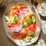 A platter with sliced watermelon, cantaloupe, cucumber, and red onion. Surrounding the platter are small bowls of feta cheese, walnuts, pepper, red pepper flakes, and a dish of honey. Sunlight casts shadows across the scene.