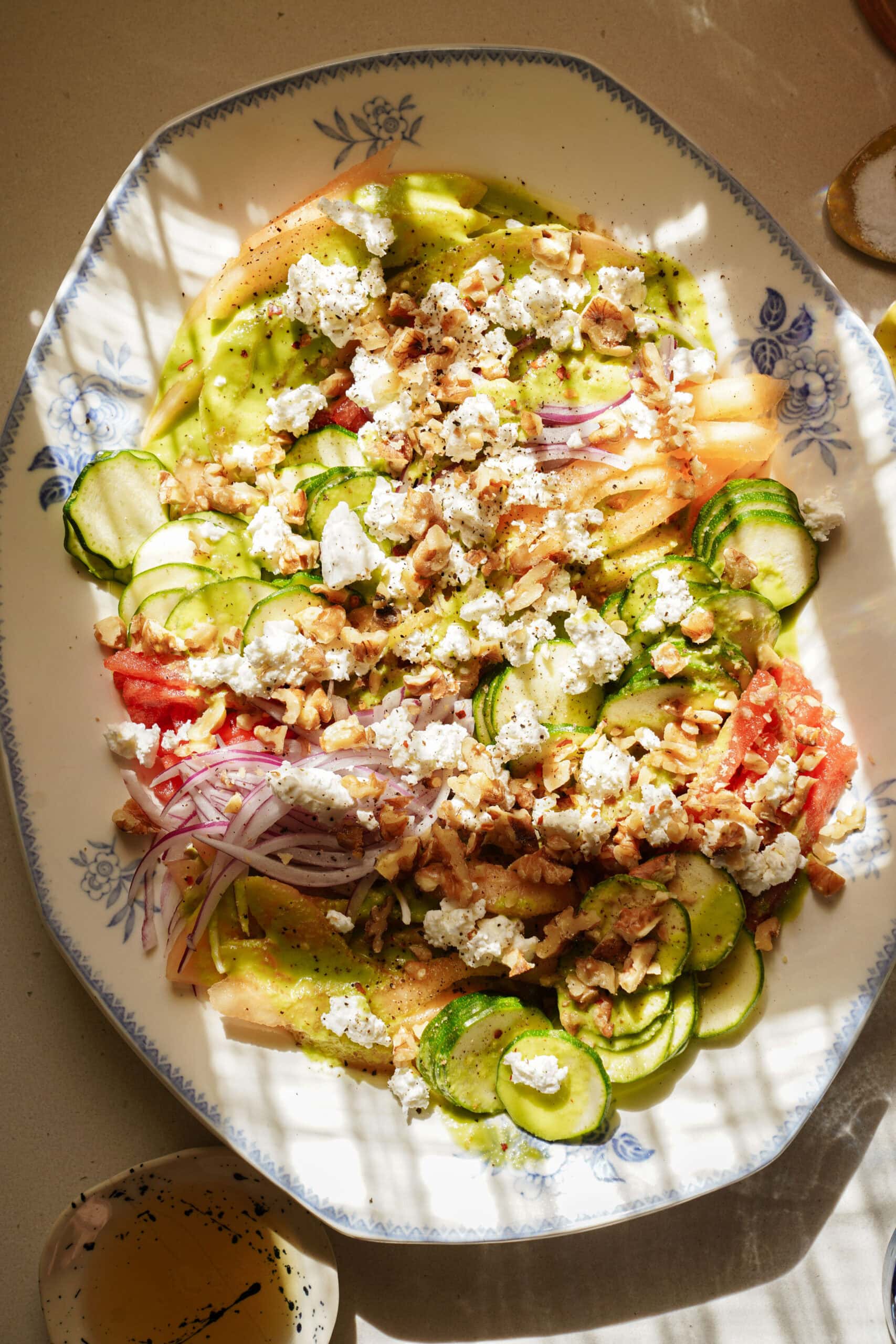 A platter with sliced cucumbers, tomatoes, onions, crumbled cheese, and chopped nuts, garnished with black pepper, sits on a sunlit table, casting shadows over the fresh, colorful salad.
