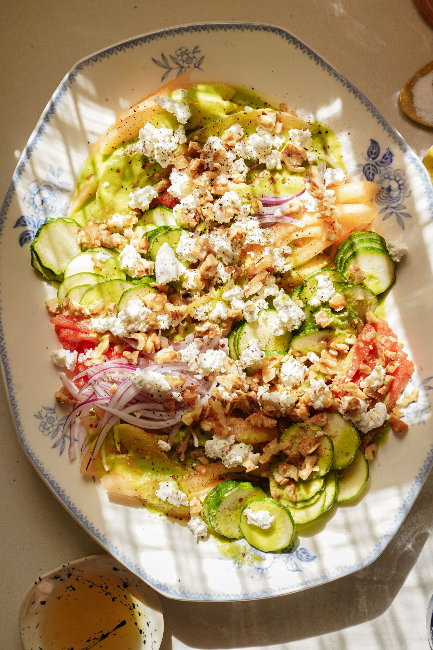 A plate of sliced cucumbers, tomatoes, onions, crumbled cheese, and chopped walnuts, drizzled with dressing, sits on a patterned dish in sunlight with shadows.