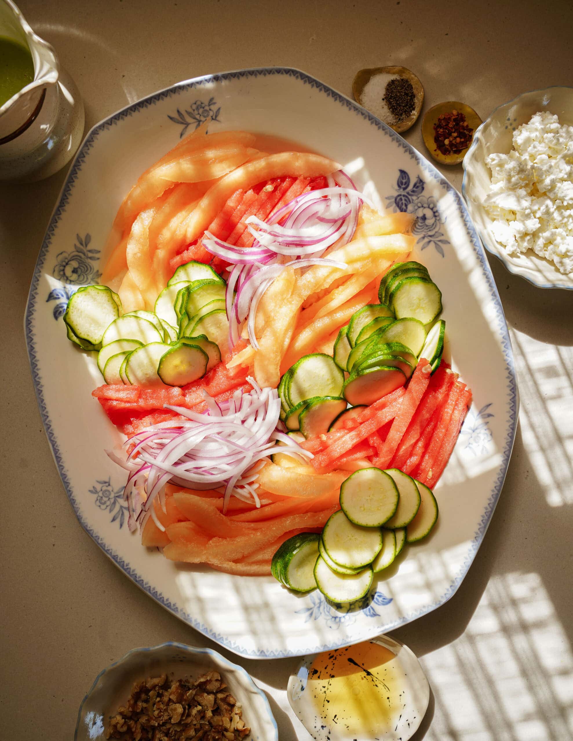 A platter with sliced watermelon, cantaloupe, cucumber, and red onion. Surrounding the platter are small bowls of feta cheese, walnuts, pepper, red pepper flakes, and a dish of honey. Sunlight casts shadows across the scene.