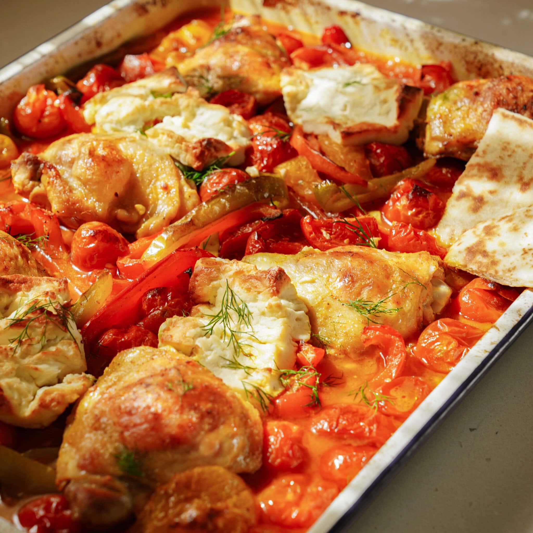 Roasted chicken thighs with golden skin, surrounded by cherry tomatoes, sliced bell peppers, and topped with dollops of white cheese, garnished with fresh herbs, served in a baking tray with a piece of flatbread.