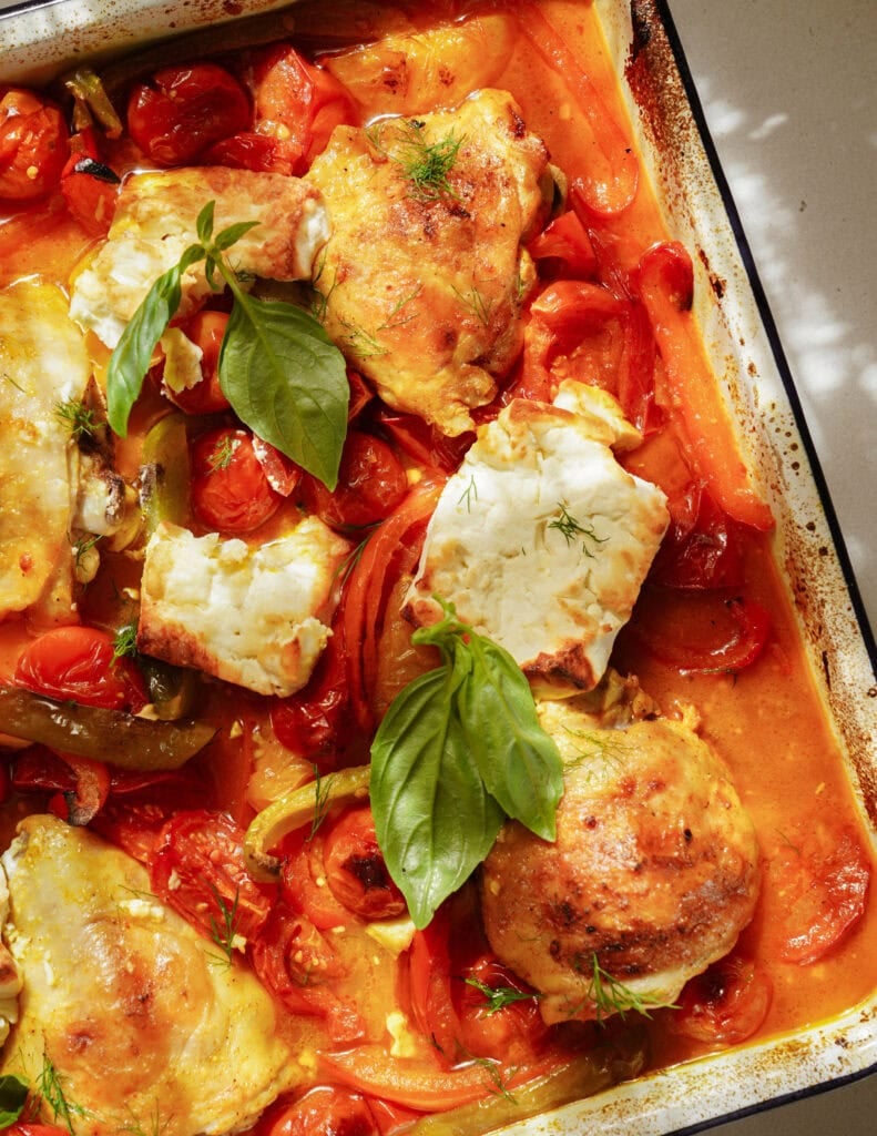 Roasted chicken thighs with tomatoes, peppers, and onions in a creamy orange sauce, topped with herbs and basil leaves, in a baking tray.