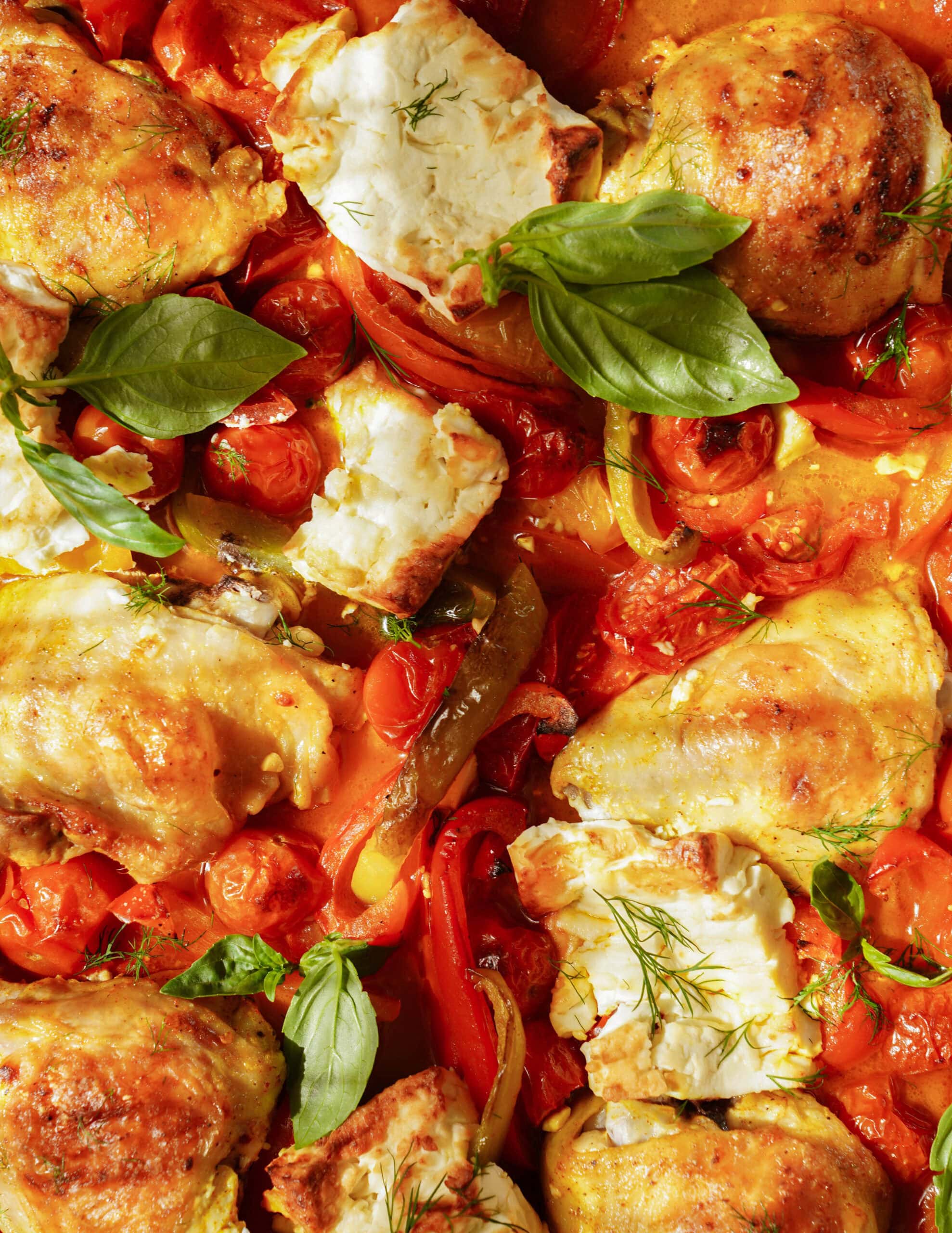 Roasted chicken thighs with tomatoes, bell peppers, chunks of white cheese, and fresh basil leaves, all baked together in a colorful, vibrant dish.