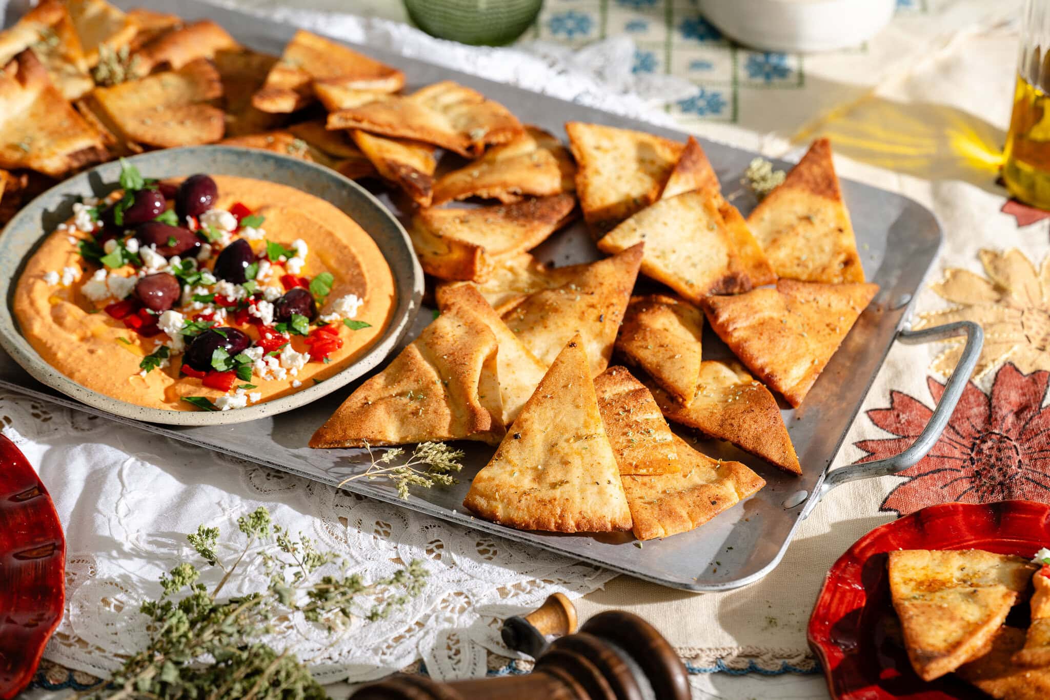 A tray of crispy pita chips is served with a plate of hummus topped with olives, feta cheese, and chopped vegetables, set on a table with sunlight streaming in.