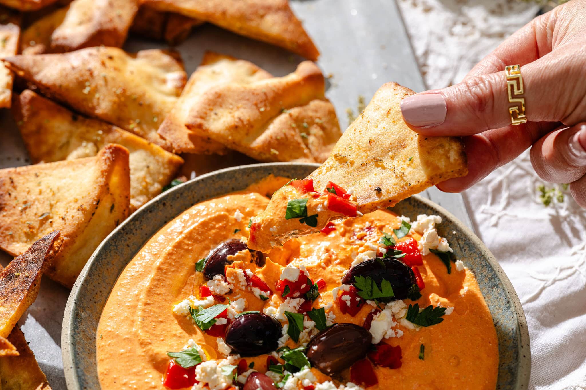 A hand dips a pita chip into a bowl of creamy dip topped with olives, feta cheese, chopped herbs, and red peppers. More pita chips are scattered on a tray in the background.