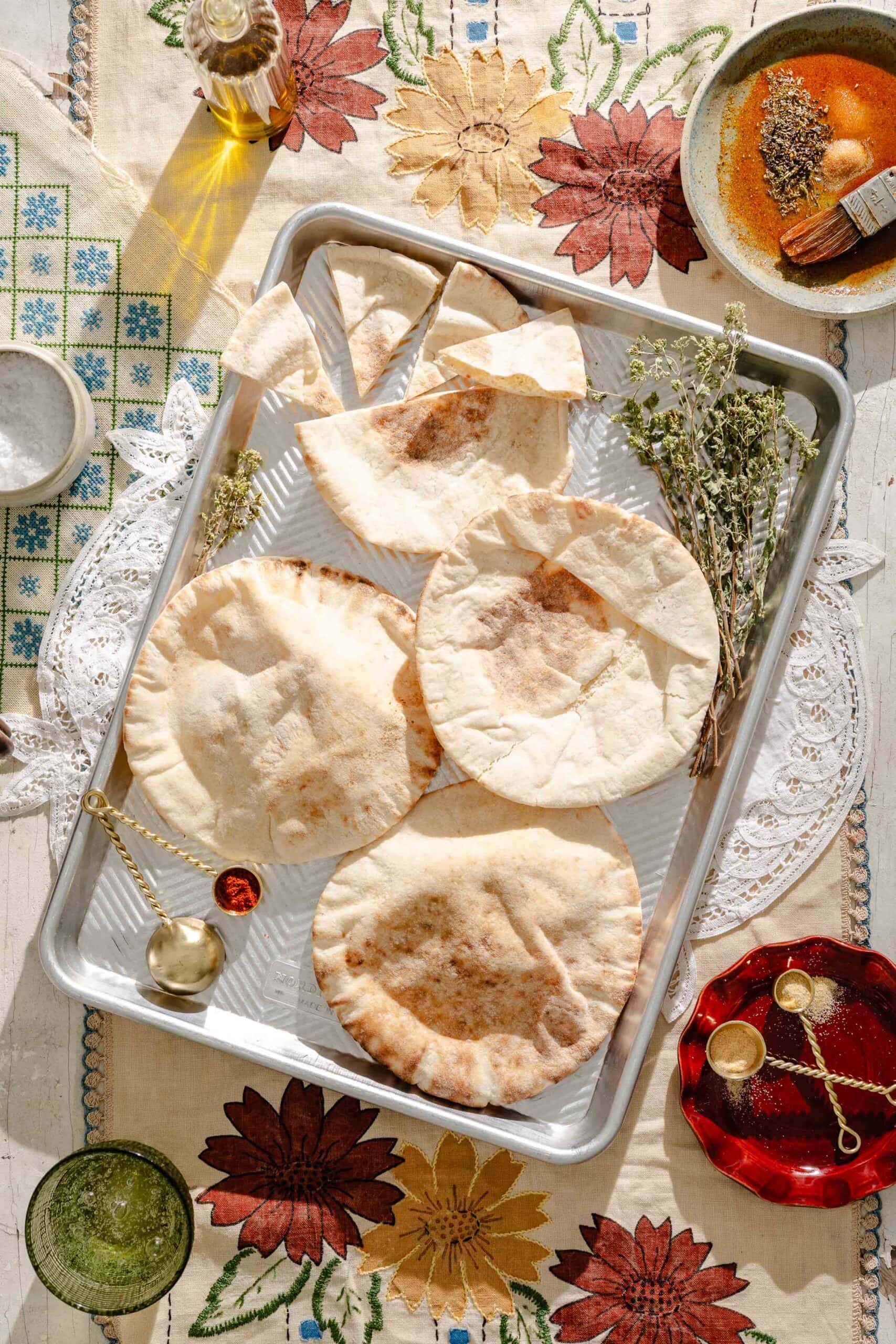 Four pieces of pita bread on a baking tray with dried herbs, surrounded by a patterned tablecloth, olive oil, a bowl with spices, and measuring spoons.