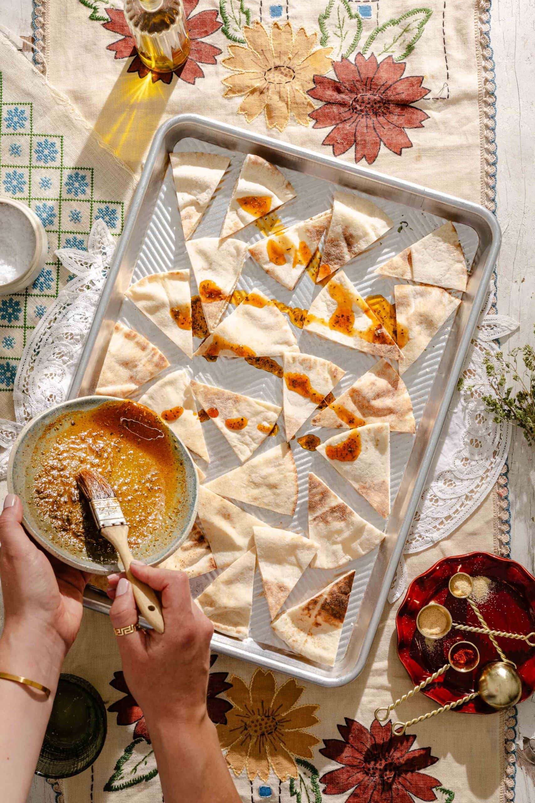 A hand brushes spiced oil onto sliced pita bread arranged on a baking tray, atop a floral tablecloth with small dishes and a bottle of olive oil nearby.