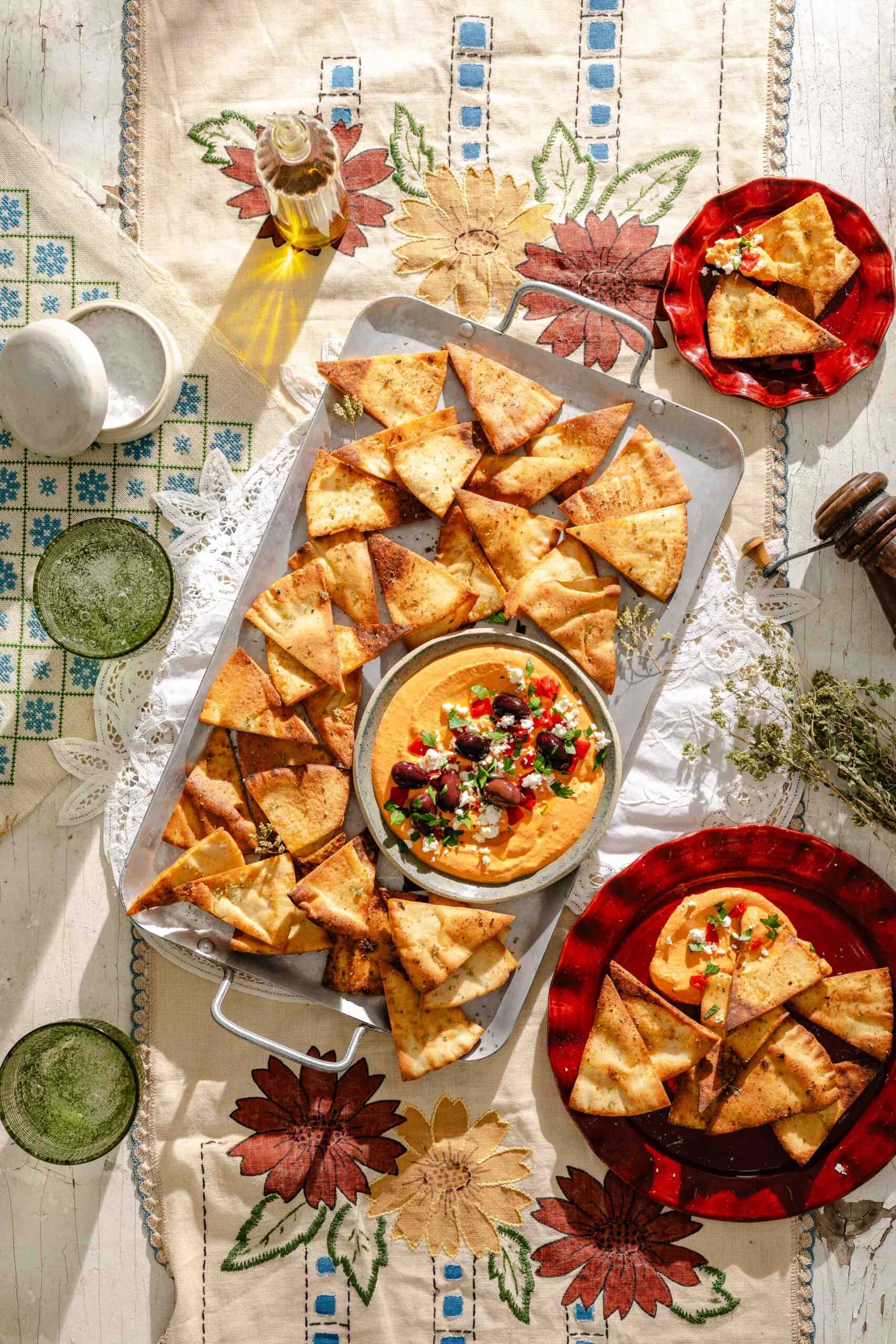 A tray of pita chips with a bowl of hummus topped with vegetables, set on a floral tablecloth with two red plates of pita chips, drinks, olive oil, and herbs on the side.