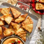 A tray of baked pita chips seasoned with herbs sits on a tablecloth, next to a bowl of hummus and a red plate with more chips and feta cheese. Sunlight highlights the crispy texture of the chips.
