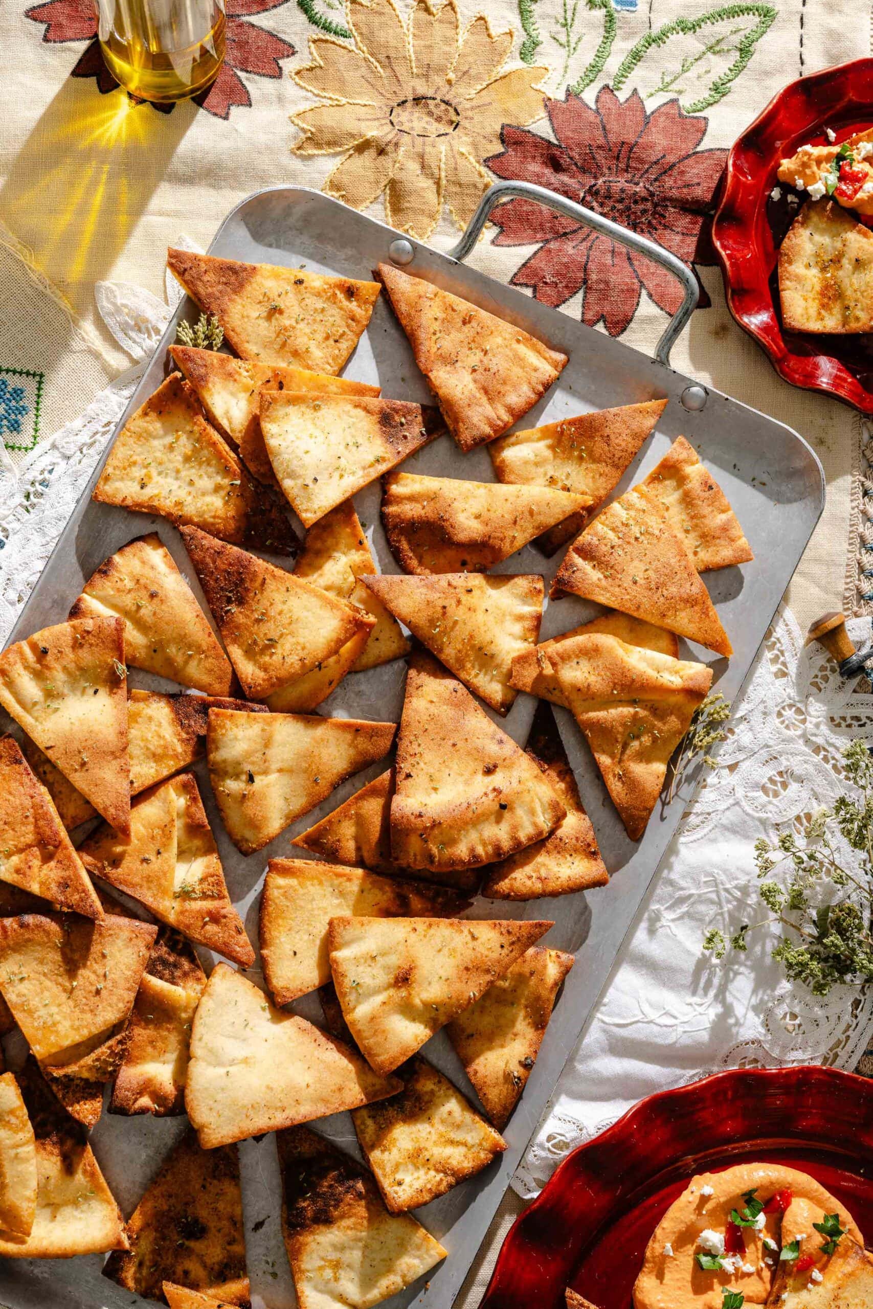 A tray of golden brown, crispy pita chips sits on a patterned tablecloth, surrounded by plates with dips and garnishes in bright sunlight.