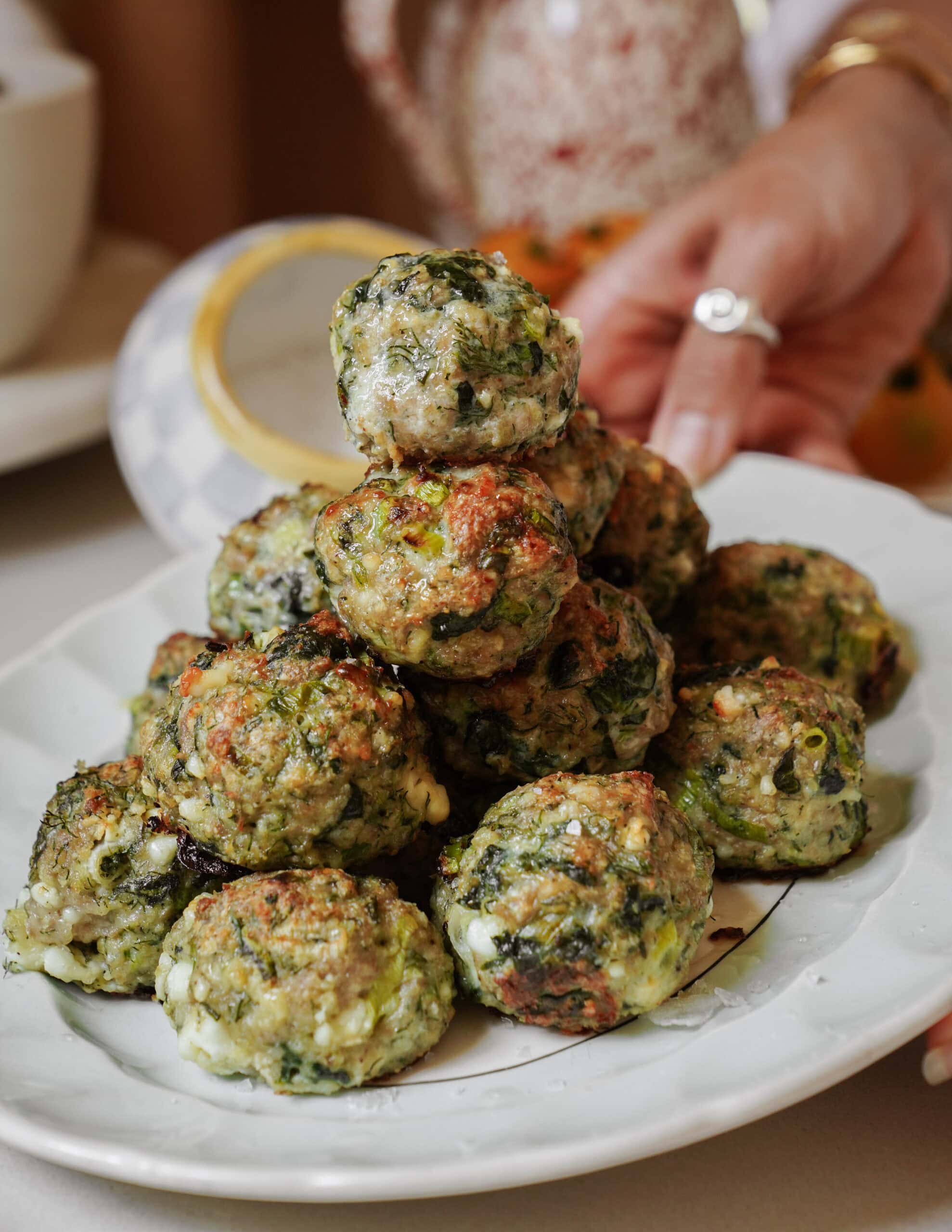 A hand reaches for a plate stacked with baked spinach and cheese balls, served on a white dish with a patterned teapot and blurred background elements.