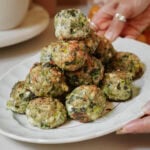 A hand holds a white plate with a stack of golden-brown spinach and cheese balls, baked and slightly crispy on the edges.
