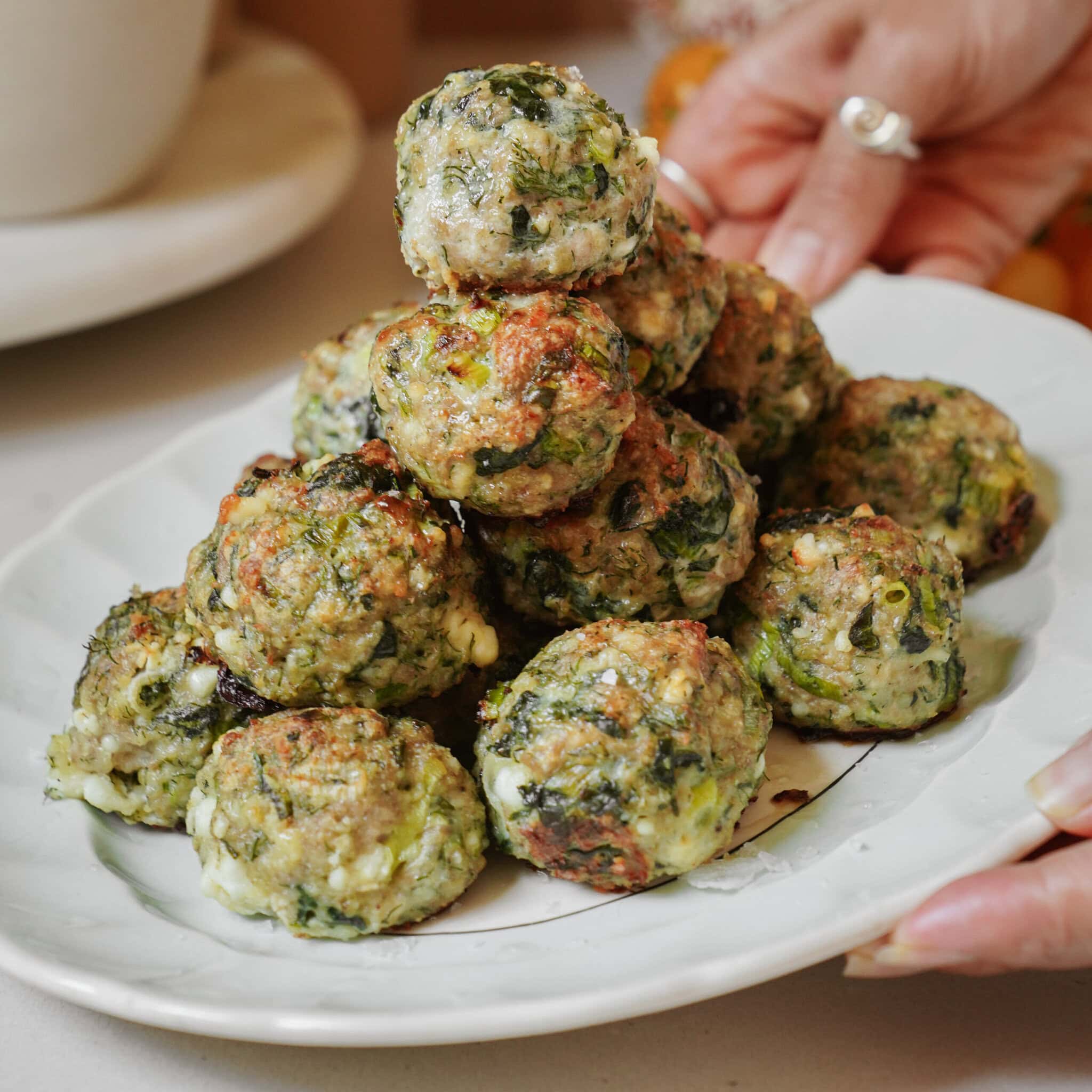 A hand holds a white plate with a stack of golden-brown spinach and cheese balls, baked and slightly crispy on the edges.