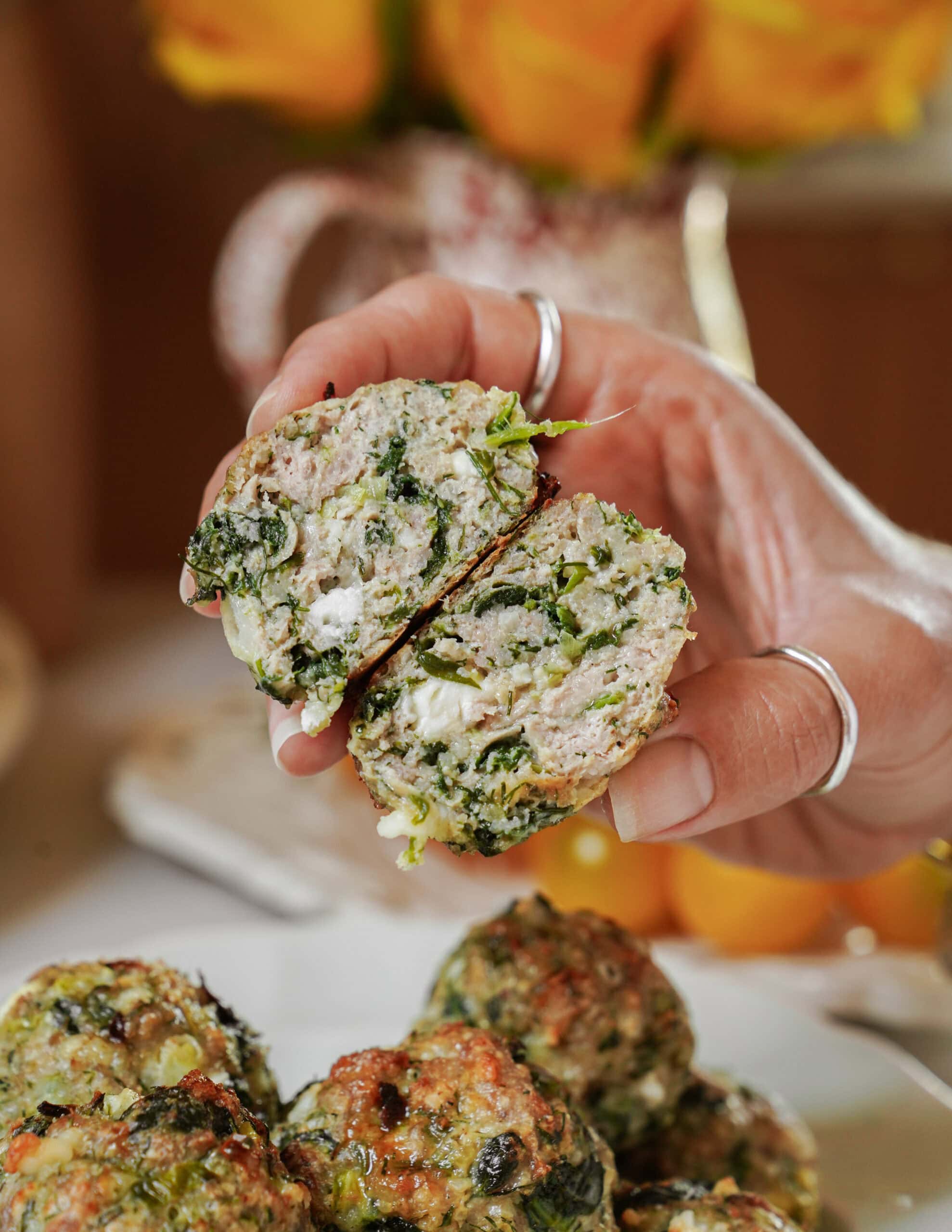 A hand holds a halved meatball filled with green herbs and white cheese. More meatballs are on a plate below. Yellow flowers are blurred in the background.